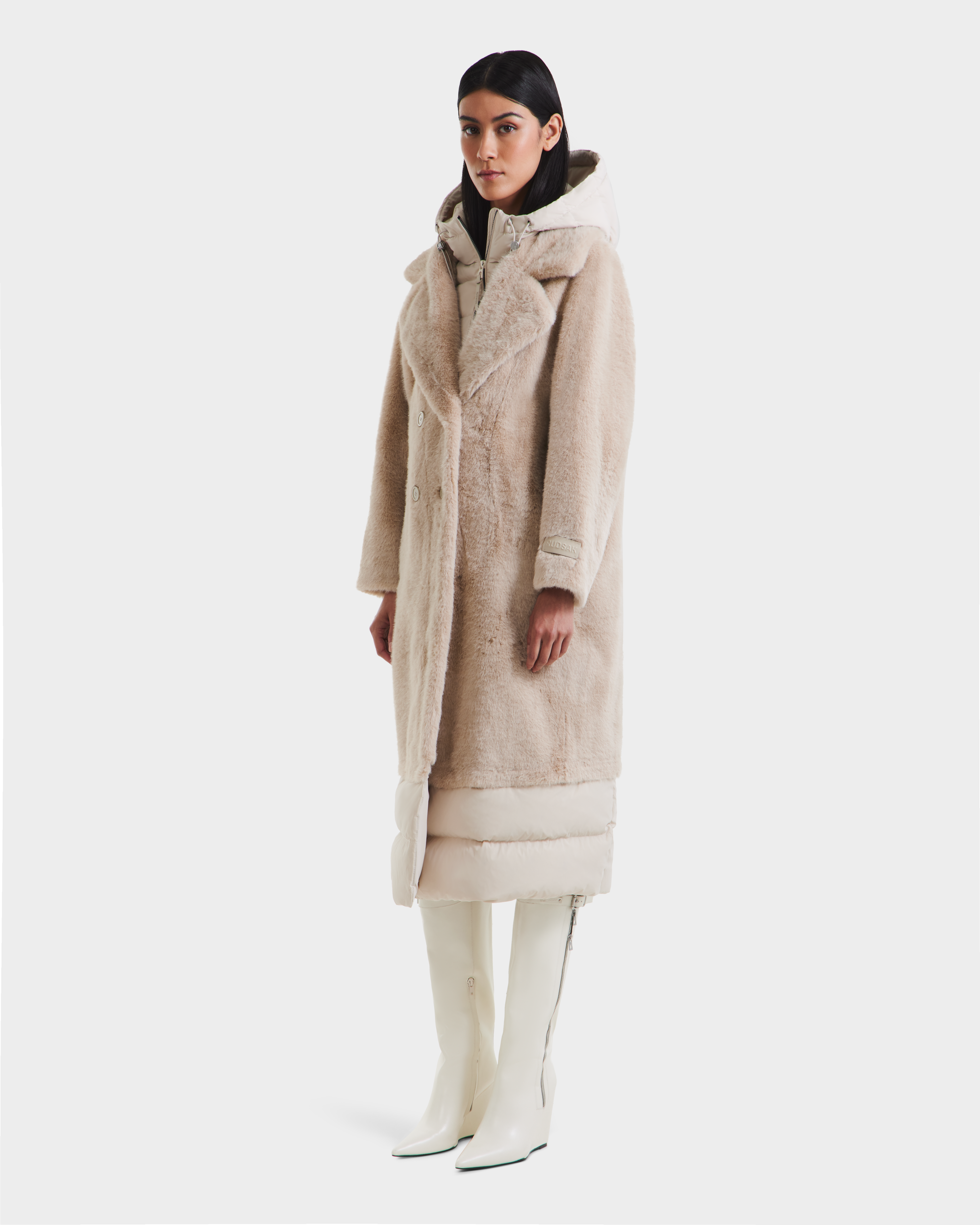 A woman wearing the Rudsak SIENA - 8125542 Vanilla faux fur coat over a white puffer, paired with white knee-high boots and a hood, poses facing forward against a plain white background.