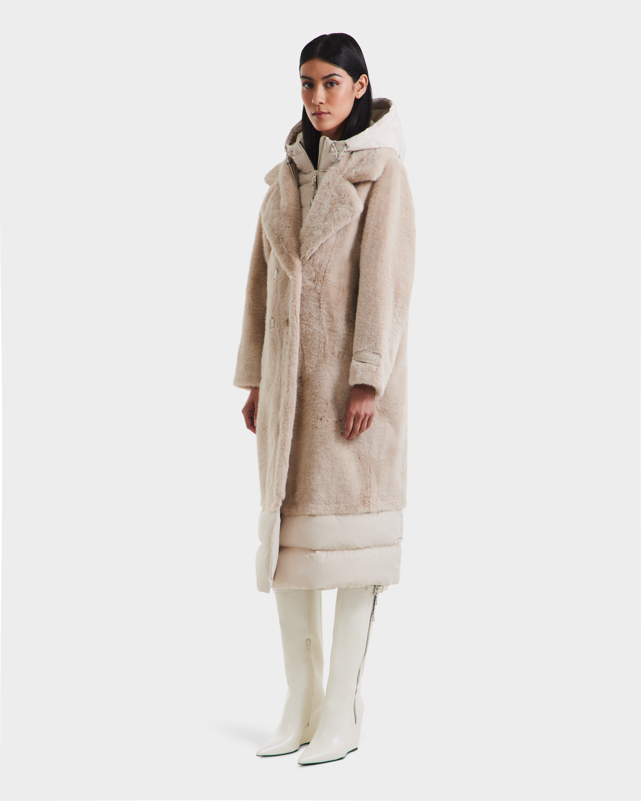 A woman wearing the Rudsak SIENA - 8125542 Vanilla faux fur coat over a white puffer, paired with white knee-high boots and a hood, poses facing forward against a plain white background.