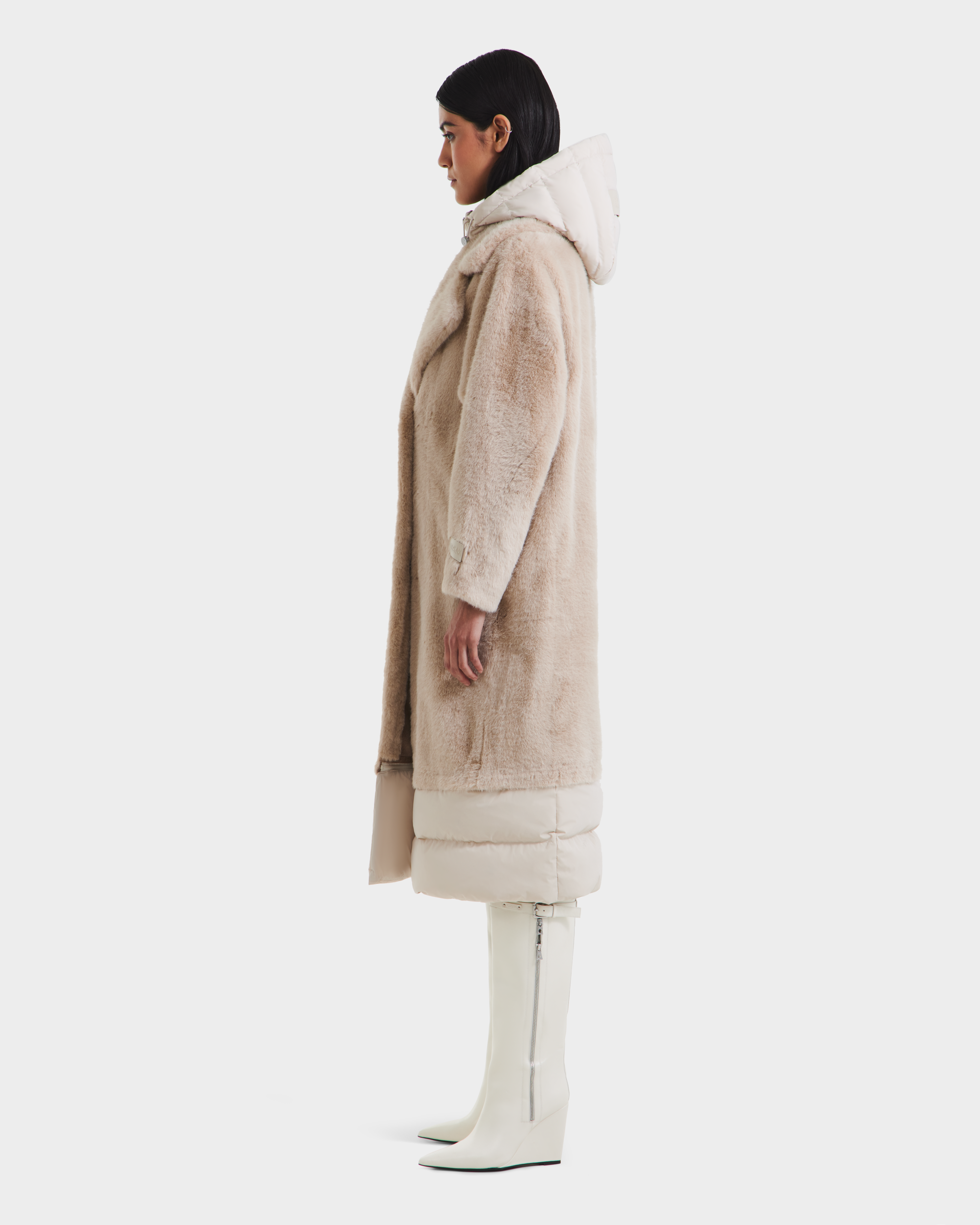 A woman stands in profile wearing the Rudsak SIENA - 8125542 Vanilla faux fur coat with a hood and light hem panel, paired with white knee-high zip boots against a plain white background.
