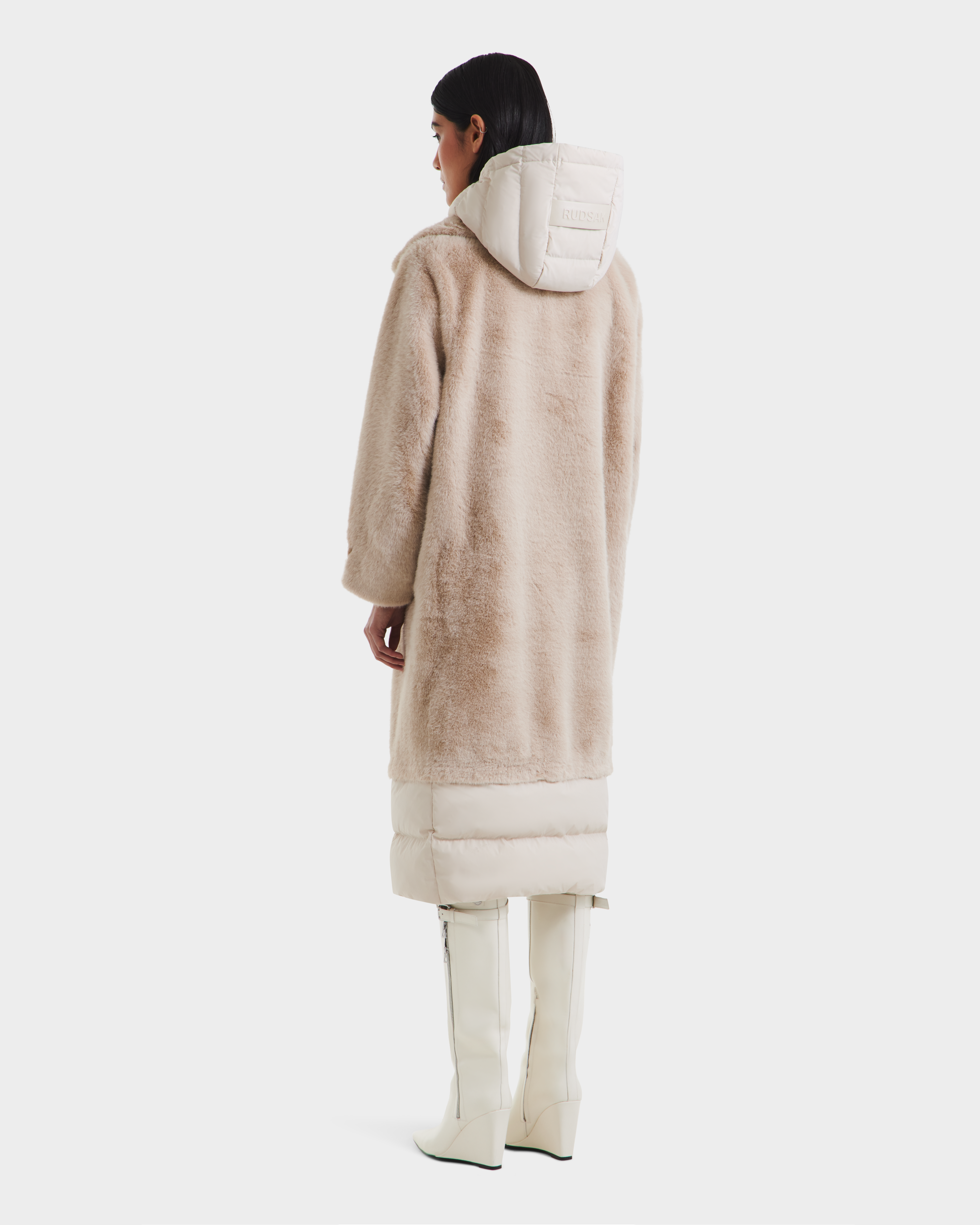 A person with straight dark hair faces away, wearing the Rudsak SIENA - 8125542 Vanilla women’s down-insulated winter coat, featuring faux fur and a quilted hood and hem, styled with white knee-high boots against a white background.