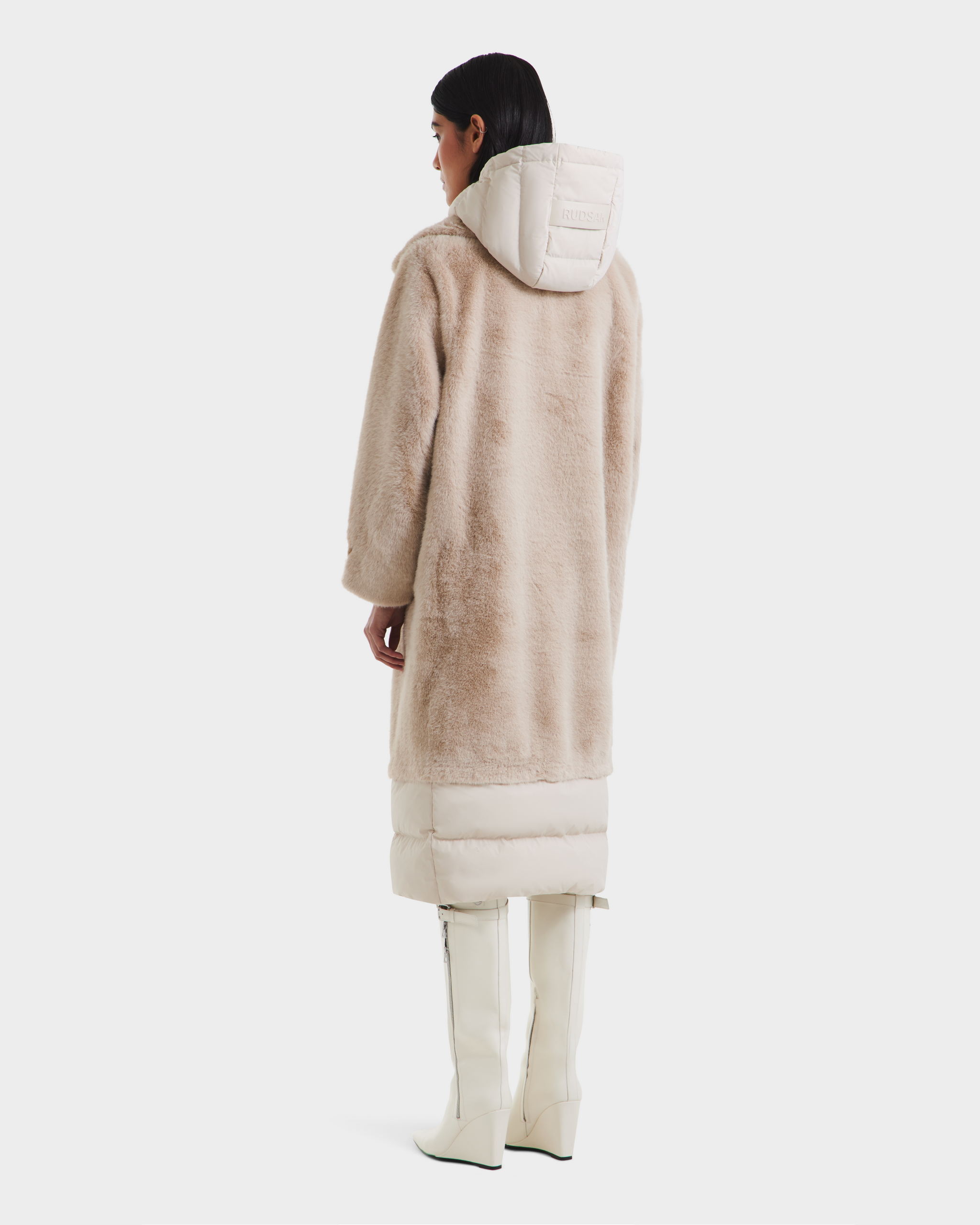 A person with straight dark hair faces away, wearing the Rudsak SIENA - 8125542 Vanilla women’s down-insulated winter coat, featuring faux fur and a quilted hood and hem, styled with white knee-high boots against a white background.