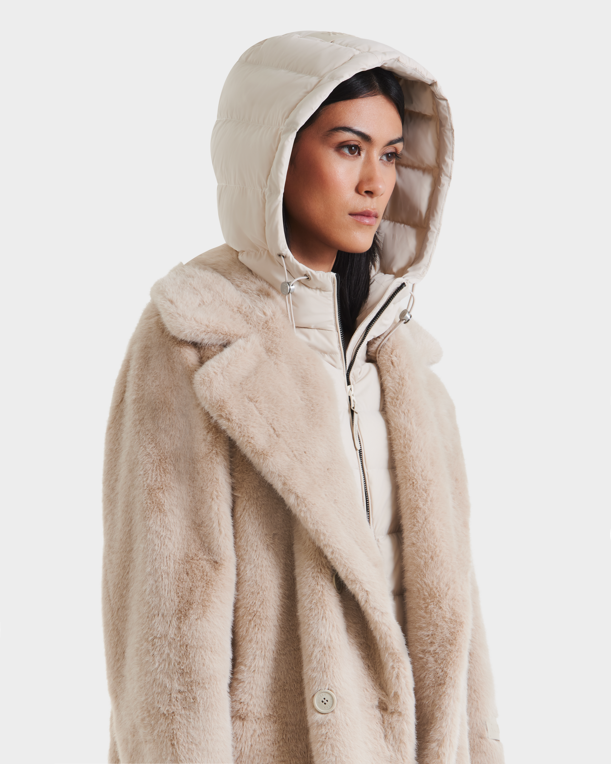 A woman wears the Rudsak SIENA - 8125542 Vanilla faux fur coat over a light down jacket with the hood up, looking to the side against a plain white background.