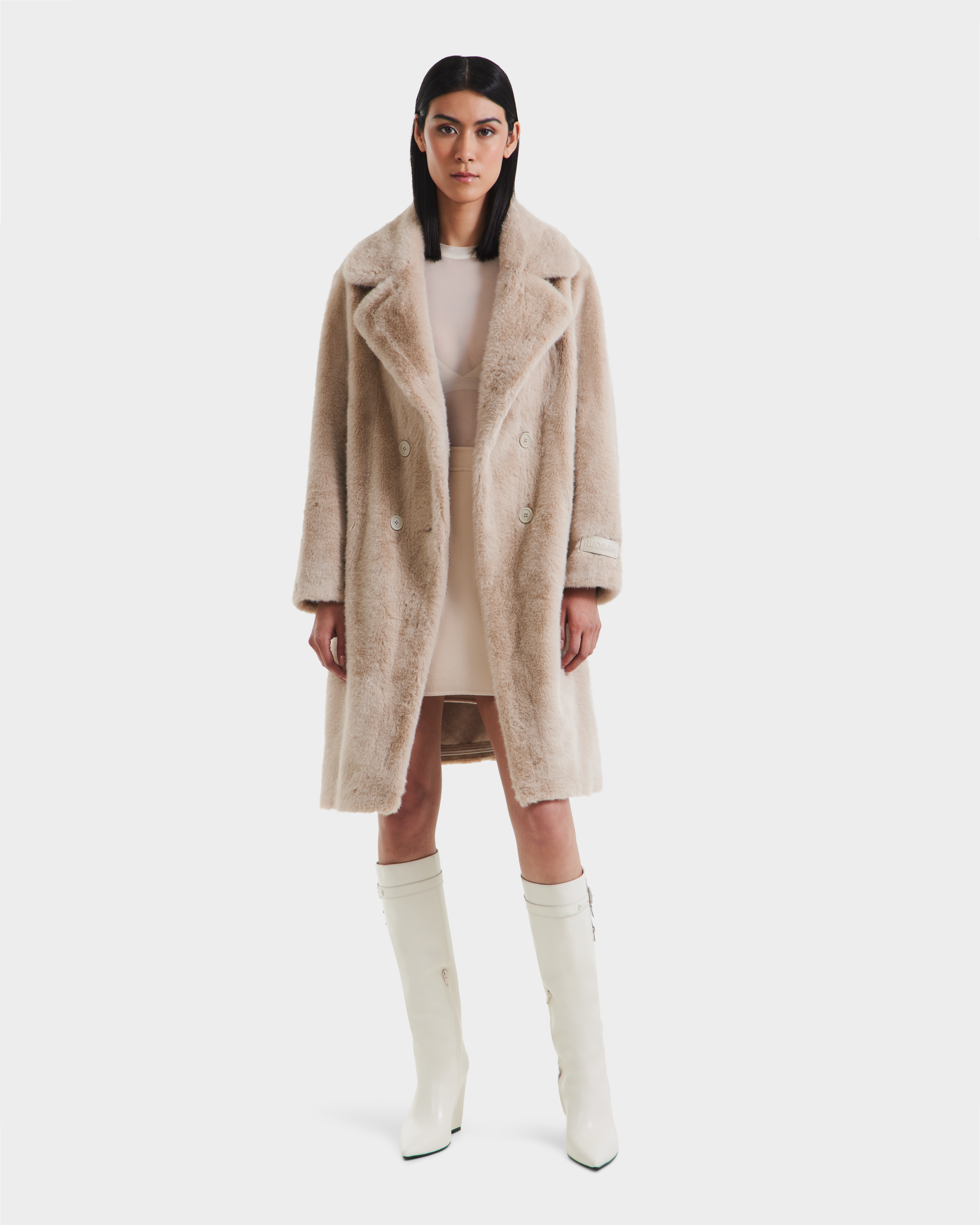 A woman with straight dark hair and a neutral expression wears the Rudsak SIENA - 8125542 Vanilla women’s faux fur coat over a short, pale dress and knee-high white boots, standing against a plain white background.