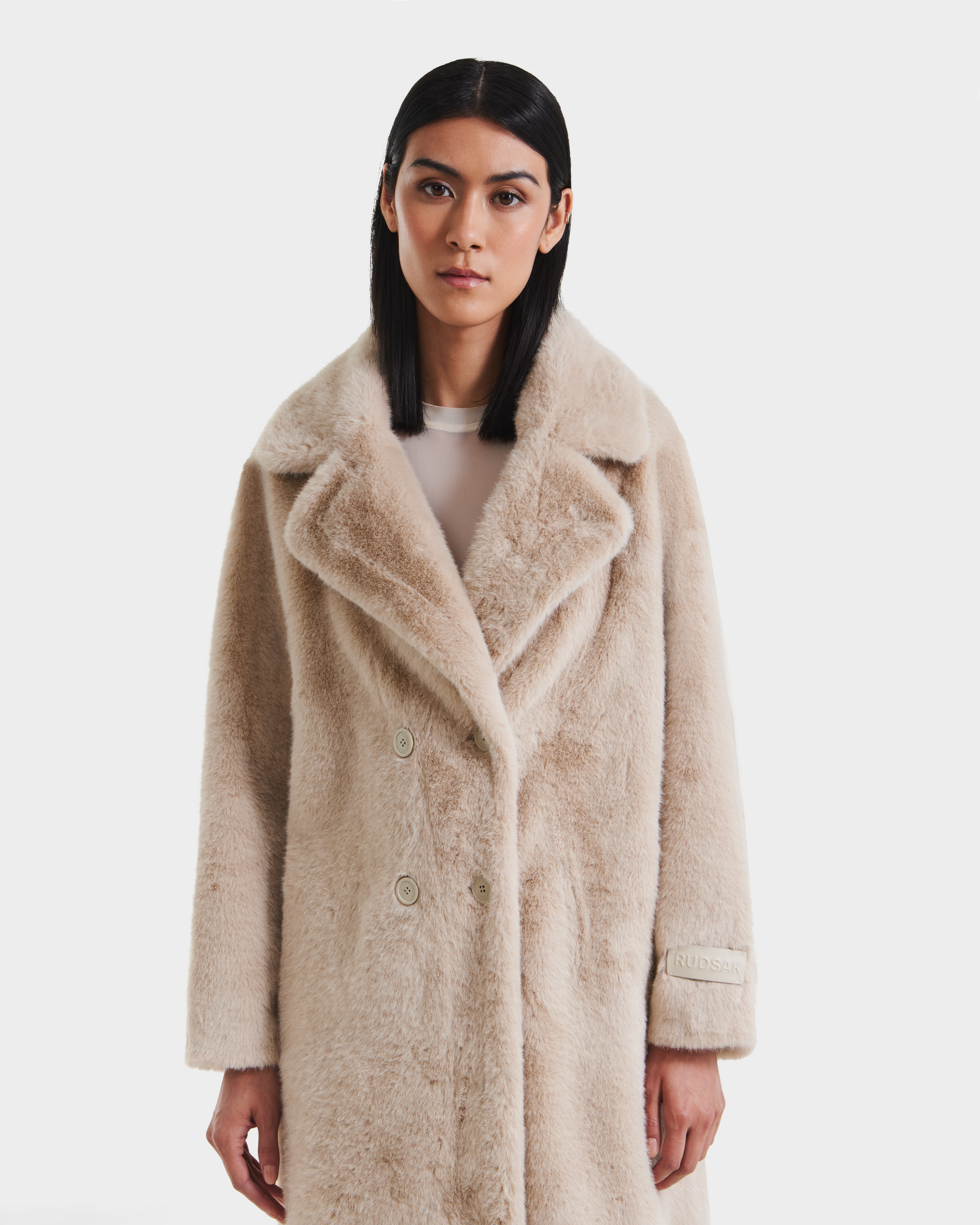A woman with straight, dark hair models the Rudsak SIENA - 8125542 Vanilla women’s faux fur coat over a light top, standing against a plain white background.