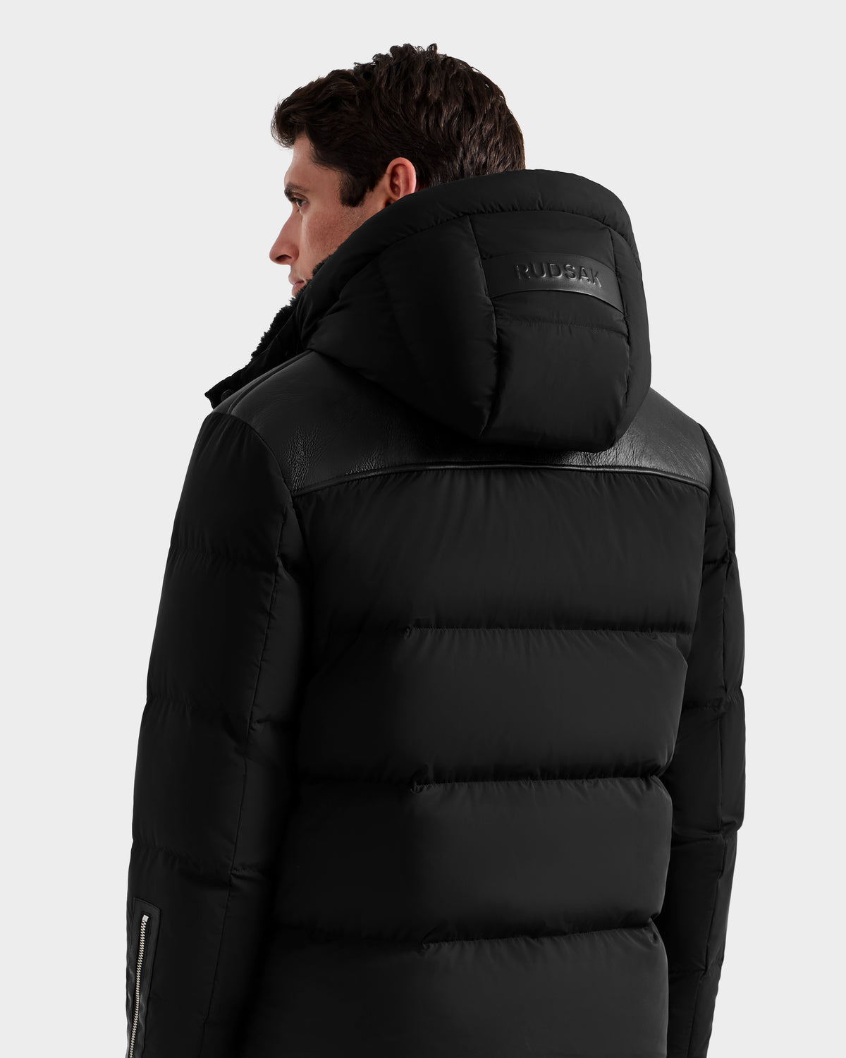 A man wearing the Rudsak SIMON - 6125597 black men's quilted down puffer with a hood, featuring leather accents and RUDSAK branding on the back of the hood, viewed from the back and slightly turned to the side.