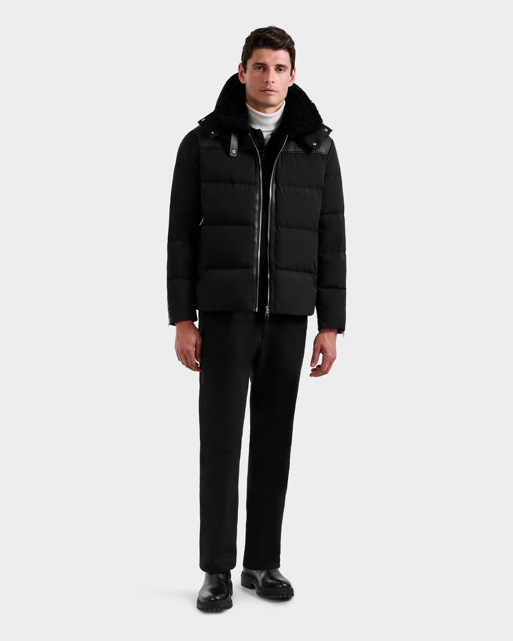 A man stands against a plain background wearing the Rudsak SIMON - 6125597 Black men's quilted down puffer with a high collar, paired with a white turtleneck, black pants, and black shoes. He looks straight at the camera with a neutral expression.