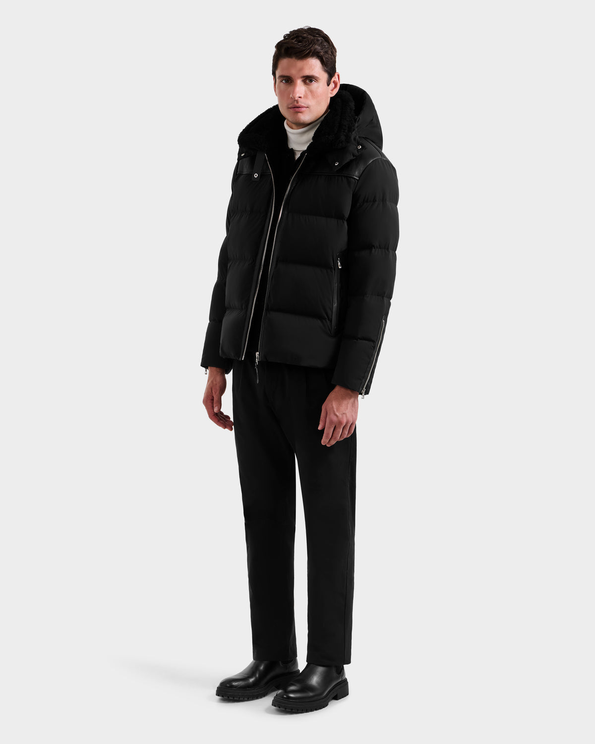 A man faces forward with a neutral expression, wearing the Rudsak SIMON - 6125597 Black men’s quilted down puffer winter jacket with a hood, paired with black pants, black boots, and a white turtleneck sweater against a plain white background.