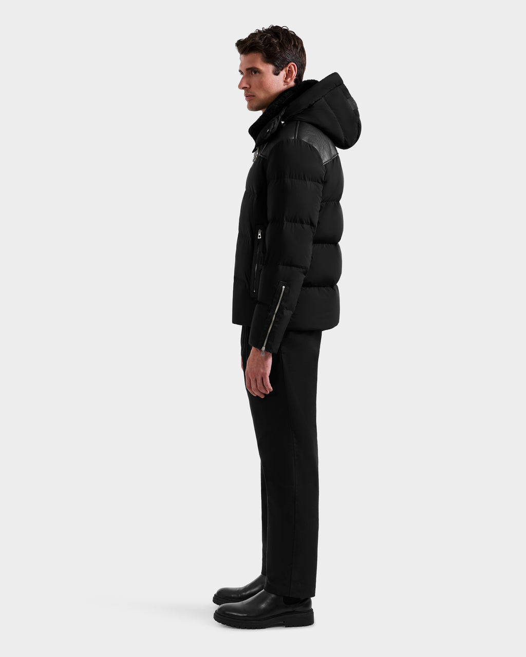 A man stands in profile against a plain background, wearing the Rudsak SIMON - 6125597 Black men’s quilted down puffer winter jacket with a hood, paired with black pants and shoes. His arms rest at his sides as he faces left.