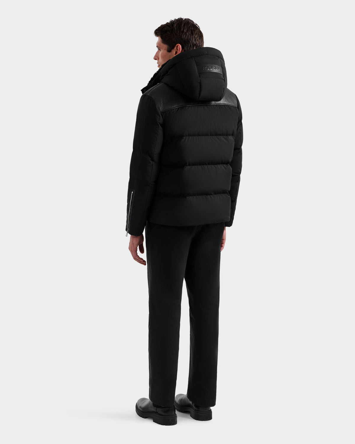 A person stands facing away, wearing the Rudsak SIMON - 6125597 Black men’s quilted down puffer with a hood, paired with black pants and shoes against a plain light gray background.