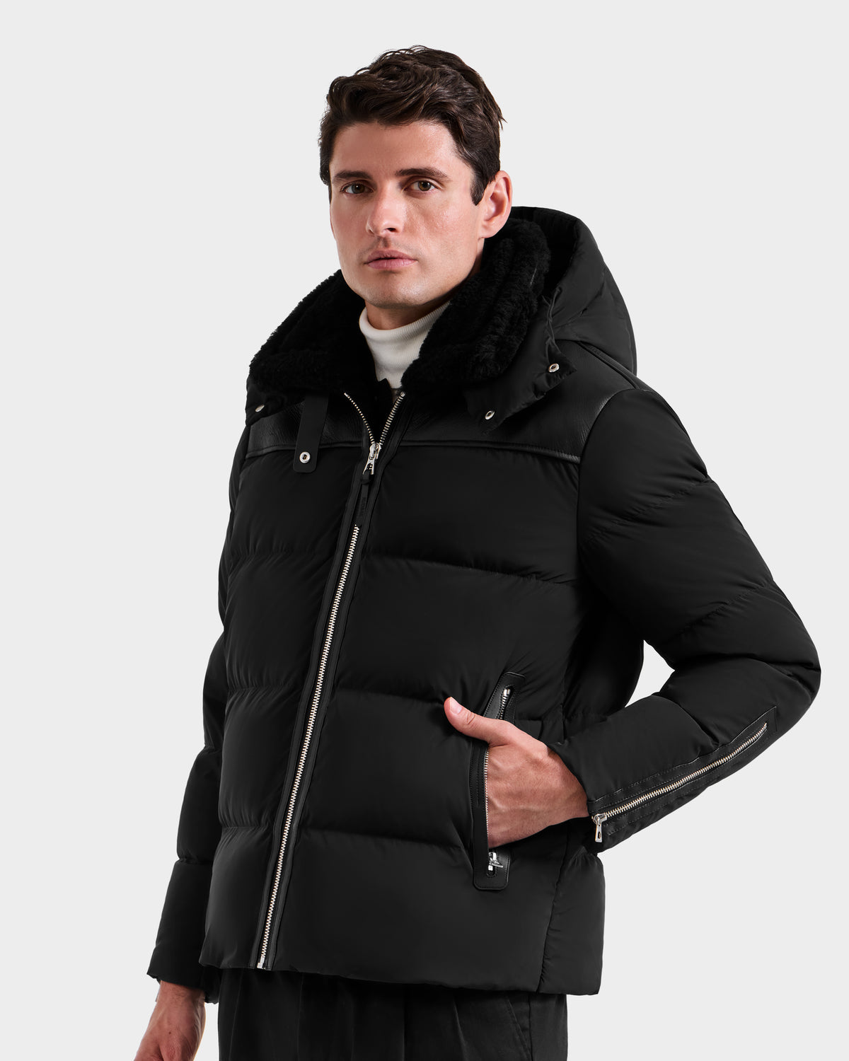 A man stands against a light gray background, wearing the Rudsak SIMON - 6125597 Black men’s down puffer jacket with a hood and zipper details, looking slightly to the side with one hand in his pocket.