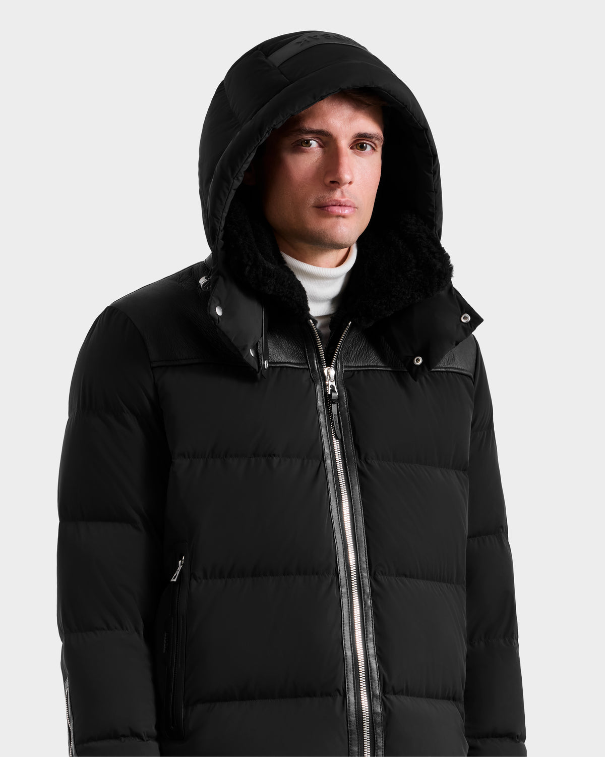 A person wearing the Rudsak SIMON - 6125597 Black men’s quilted down puffer with a hood and a white turtleneck, standing against a plain light gray background.
