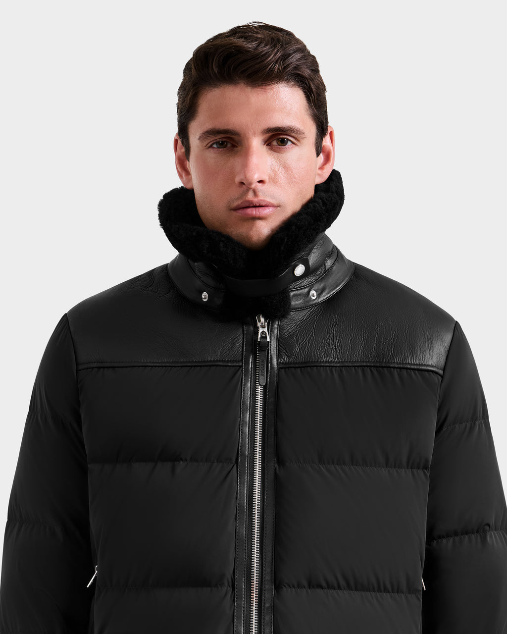 A man with short dark hair wears the Rudsak SIMON - 6125597 Black down puffer jacket with a high shearling collar, facing forward and looking at the camera against a plain light gray background.