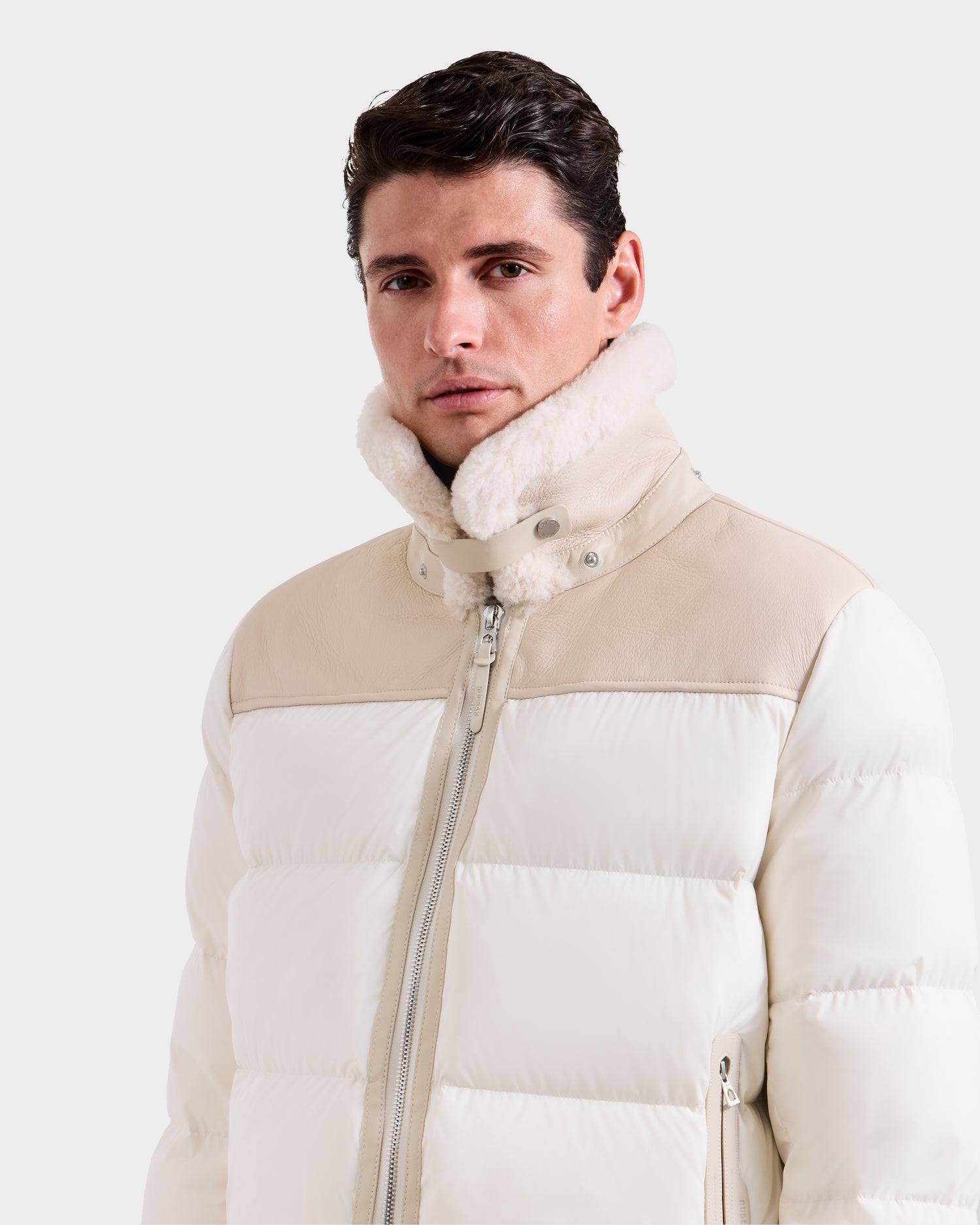 A man wearing the Rudsak SIMON - 6125597 Vanilla men's quilted down puffer jacket with a high collar and beige accents stands against a plain light background, looking slightly to the side.