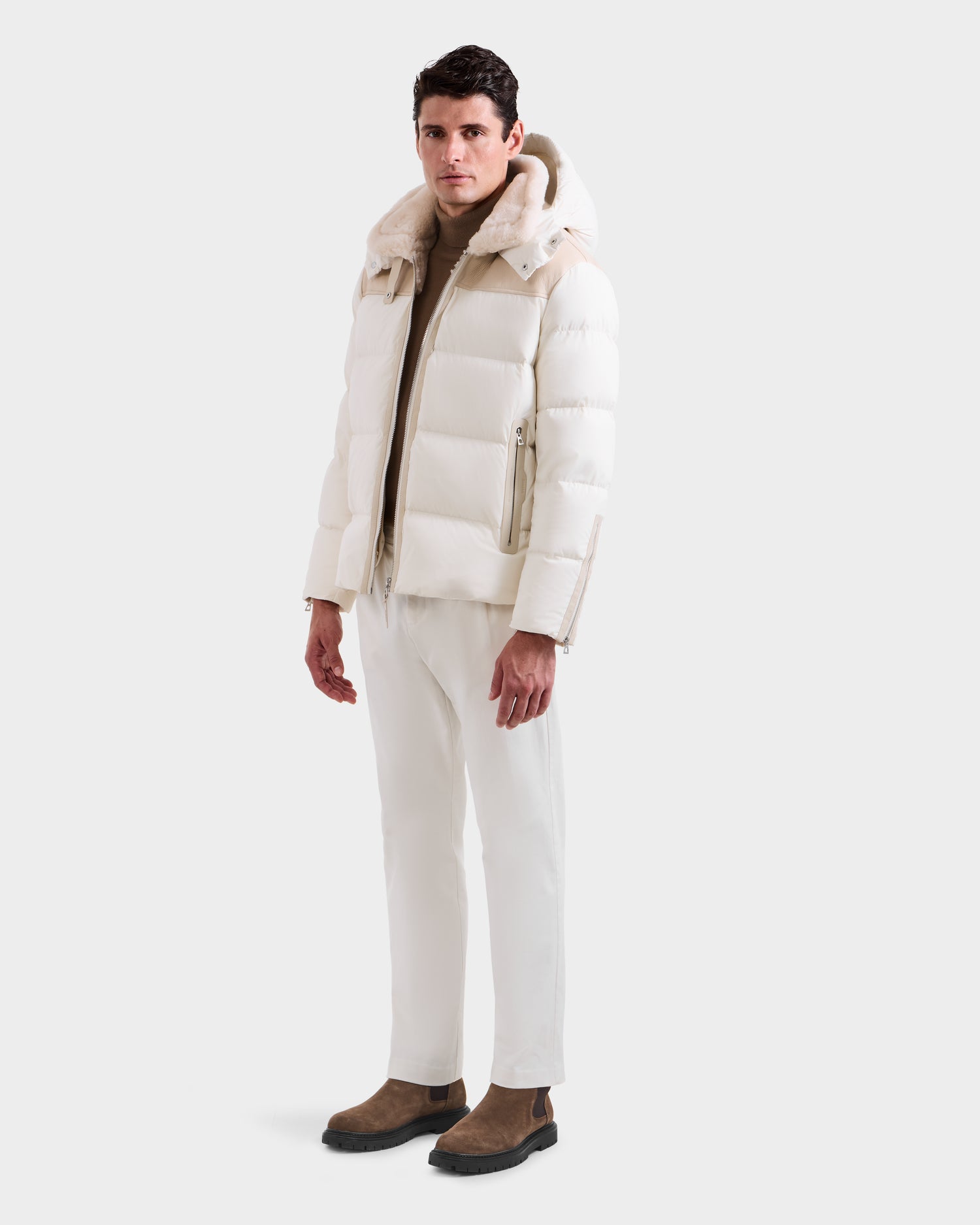 A man wears the Rudsak SIMON - 6125597 Vanilla men’s quilted down puffer jacket with a hood, paired with white pants, a beige turtleneck, and brown boots against a plain white background.