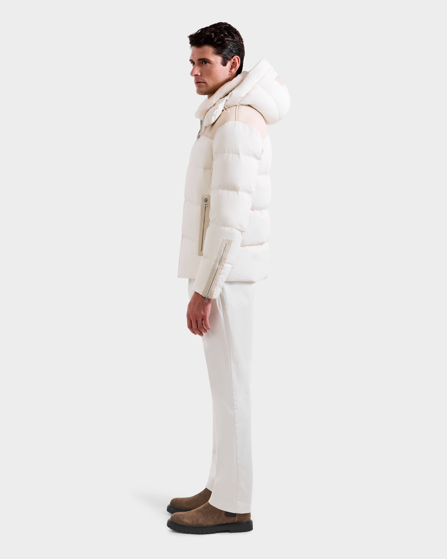 A man stands in profile against a plain light gray background wearing the Rudsak SIMON - 6125597 Vanilla men’s quilted down puffer with a hood, paired with white pants and brown suede boots.