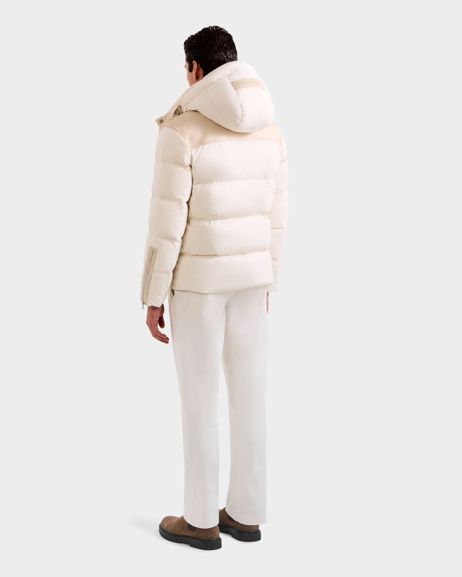 A person stands facing away against a plain light gray background, wearing Rudsak’s SIMON - 6125597 Vanilla men’s quilted down puffer jacket with a hood, paired with white pants and brown shoes.