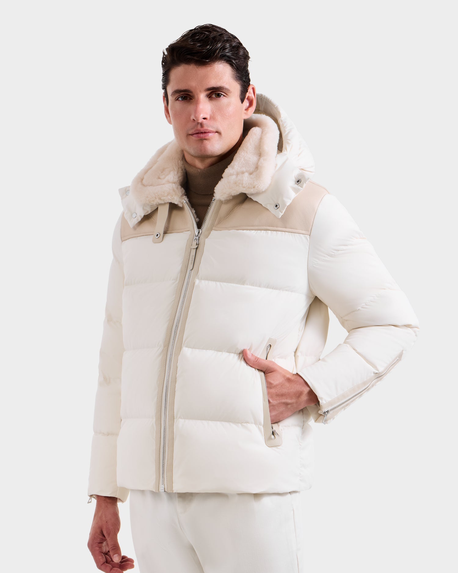 A man wears the Rudsak SIMON - 6125597 Vanilla cream down puffer jacket with a shearling collar and beige accents over a light brown turtleneck, standing with one hand in his pocket against a white background.