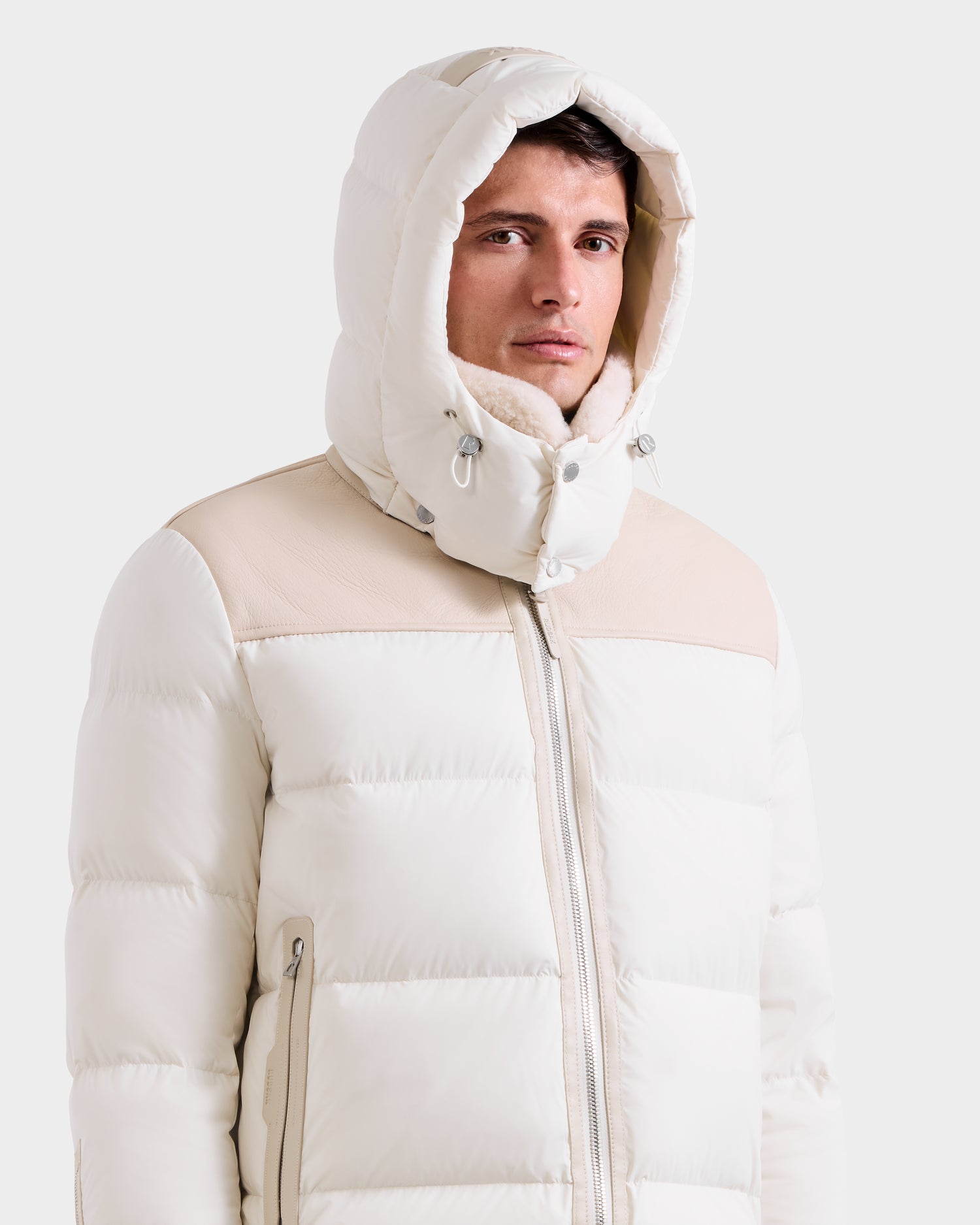 A man wearing the Rudsak SIMON - 6125597 Vanilla men’s quilted down puffer with beige accents stands against a plain white background, looking slightly to the side.