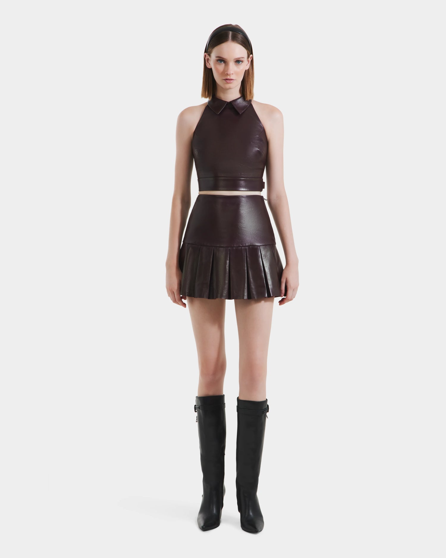 A woman faces forward in a dark brown sleeveless leather crop top with a collar, a Rudsak SOLANGE - 8524543 Bordeaux womens pleated leather skirt, and knee-high black boots against a plain white background.