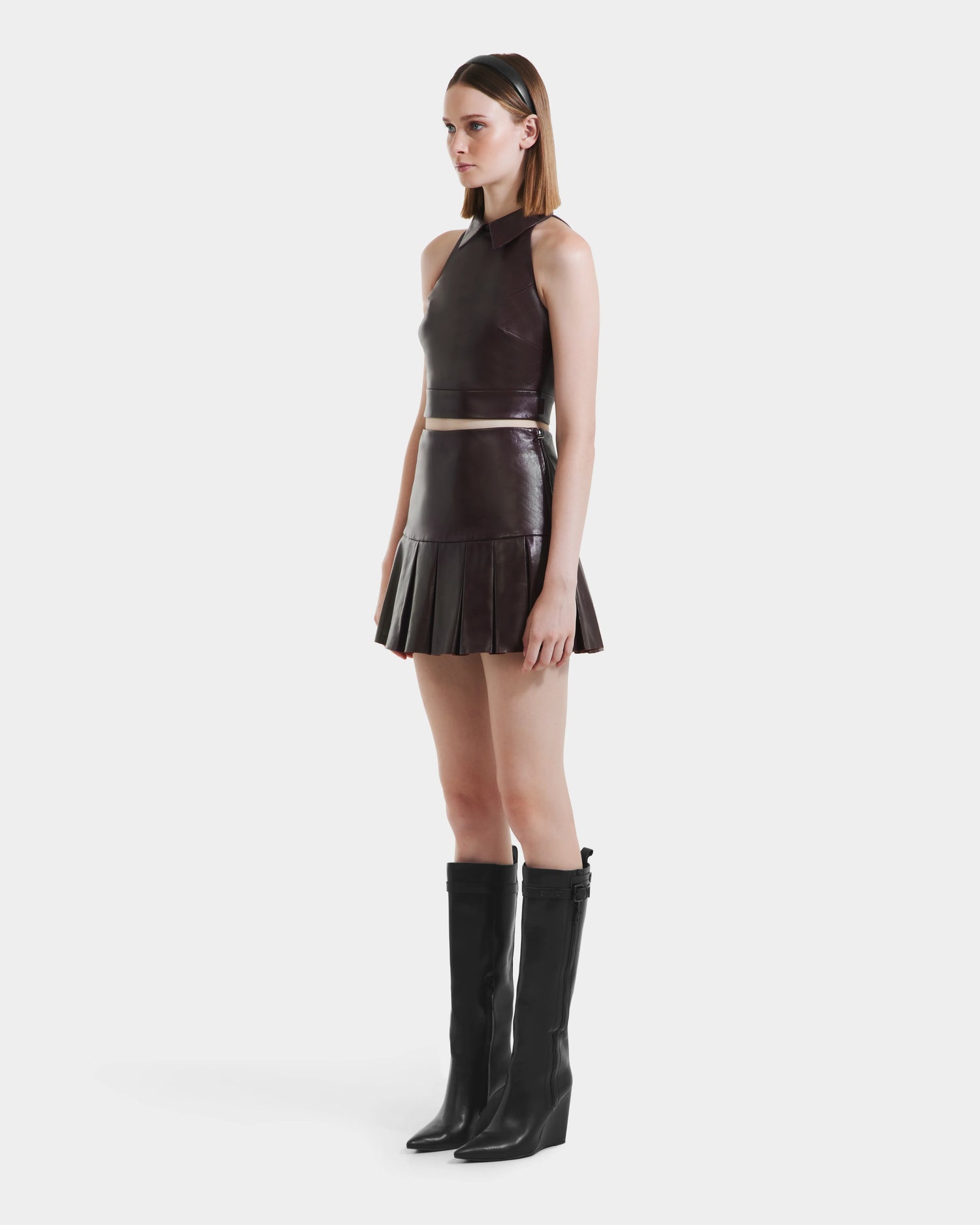 A woman in profile wears the Rudsak SOLANGE - 8524543 Bordeaux sleeveless leather top, a pleated leather skirt, and knee-high black boots. She has straight brown hair with a headband, posing against a plain white background.