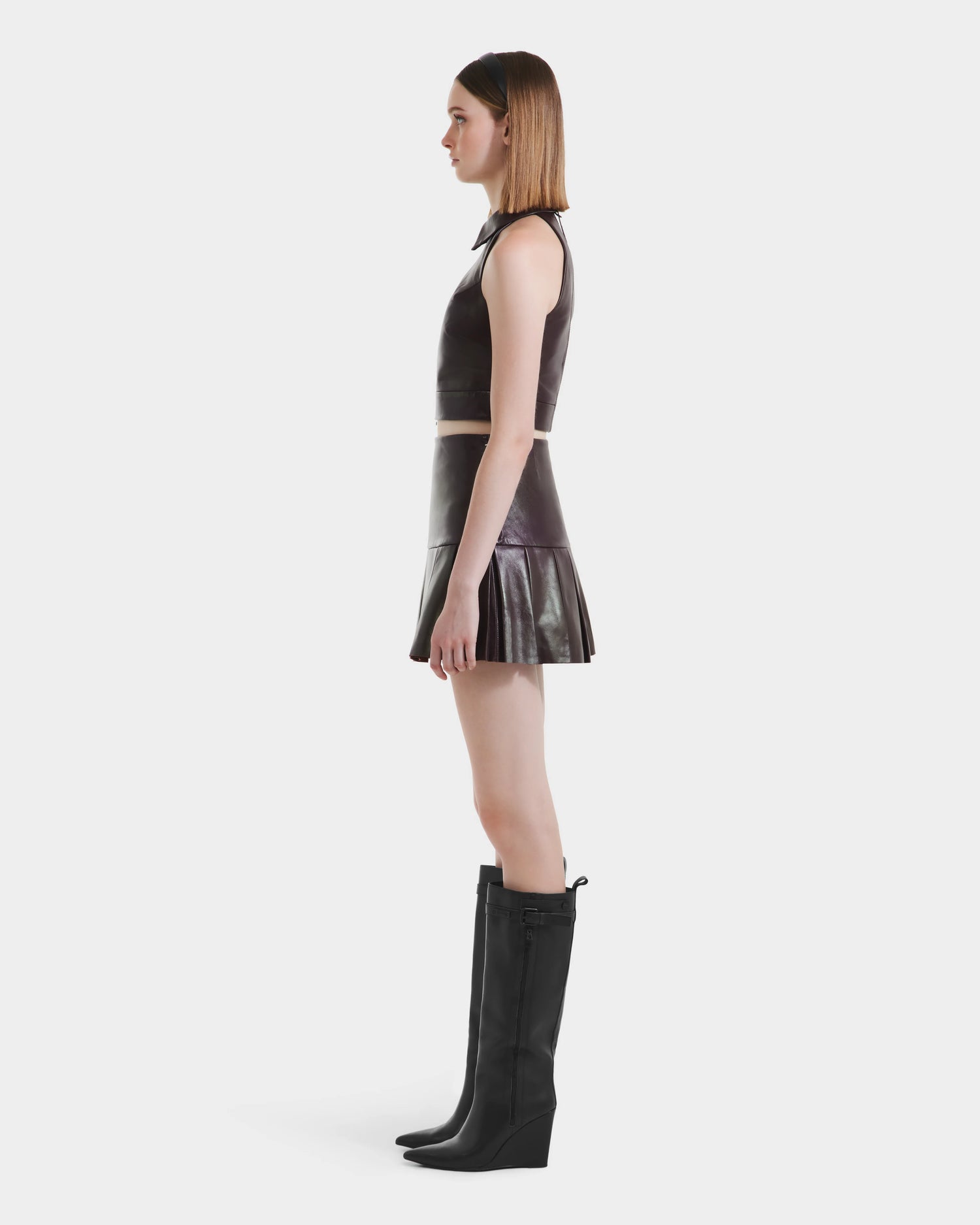 A woman stands in profile against a plain white background, wearing a sleeveless dark crop top, the Rudsak SOLANGE - 8524543 Bordeaux short leather skirt, and knee-high black wedge boots.
