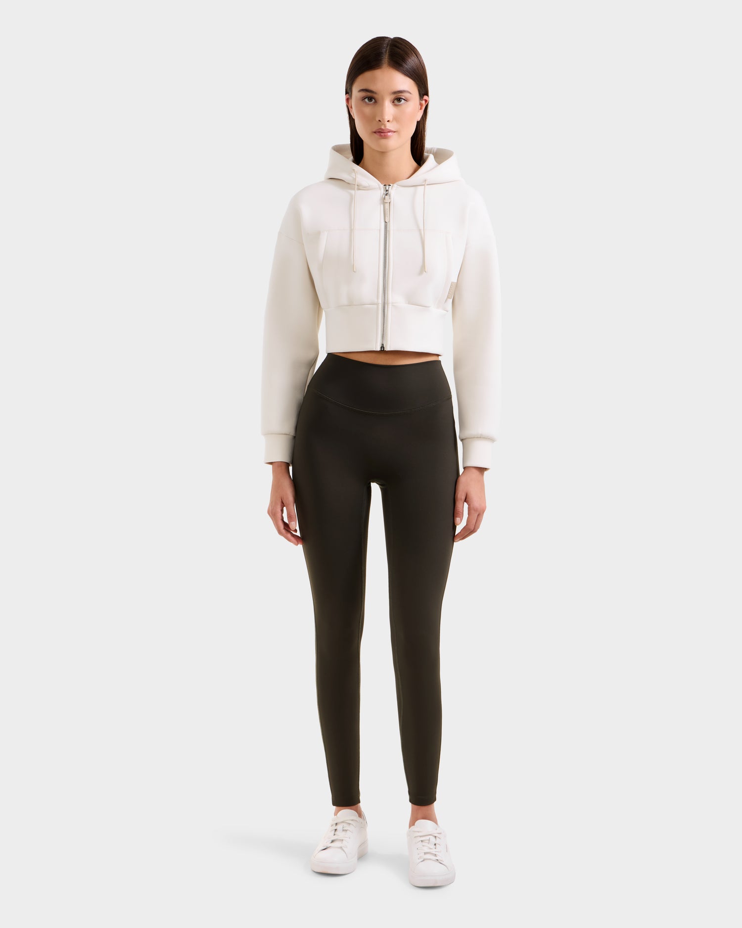 A woman stands facing forward, wearing a white cropped zip-up hoodie, Rudsak SONOMA - 8525071 Dark Olive leggings, and white sneakers against a plain light gray background.