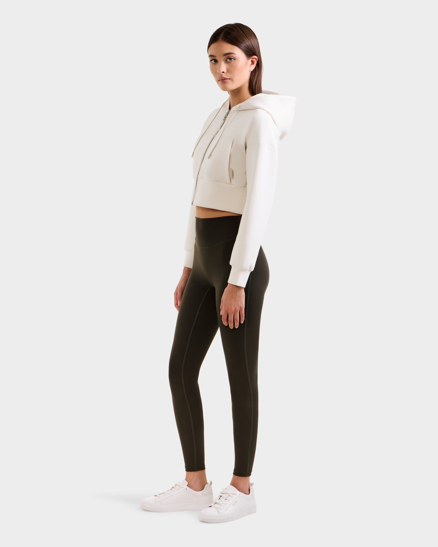 A woman stands slightly sideways in a cropped white hoodie, white sneakers, and Rudsak SONOMA - 8525071 Dark Olive high-waisted leggings against a light background—a stylish, versatile activewear look.