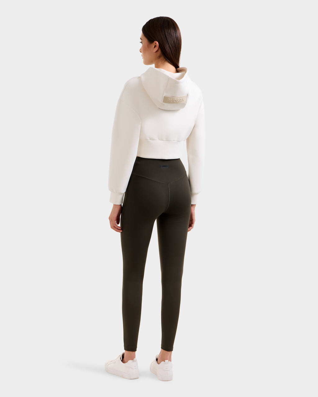 A woman faces away, wearing a cropped white hoodie with a hood and high-waisted Rudsak SONOMA - 8525071 Dark Olive leggings, paired with white sneakers, against a plain light background.