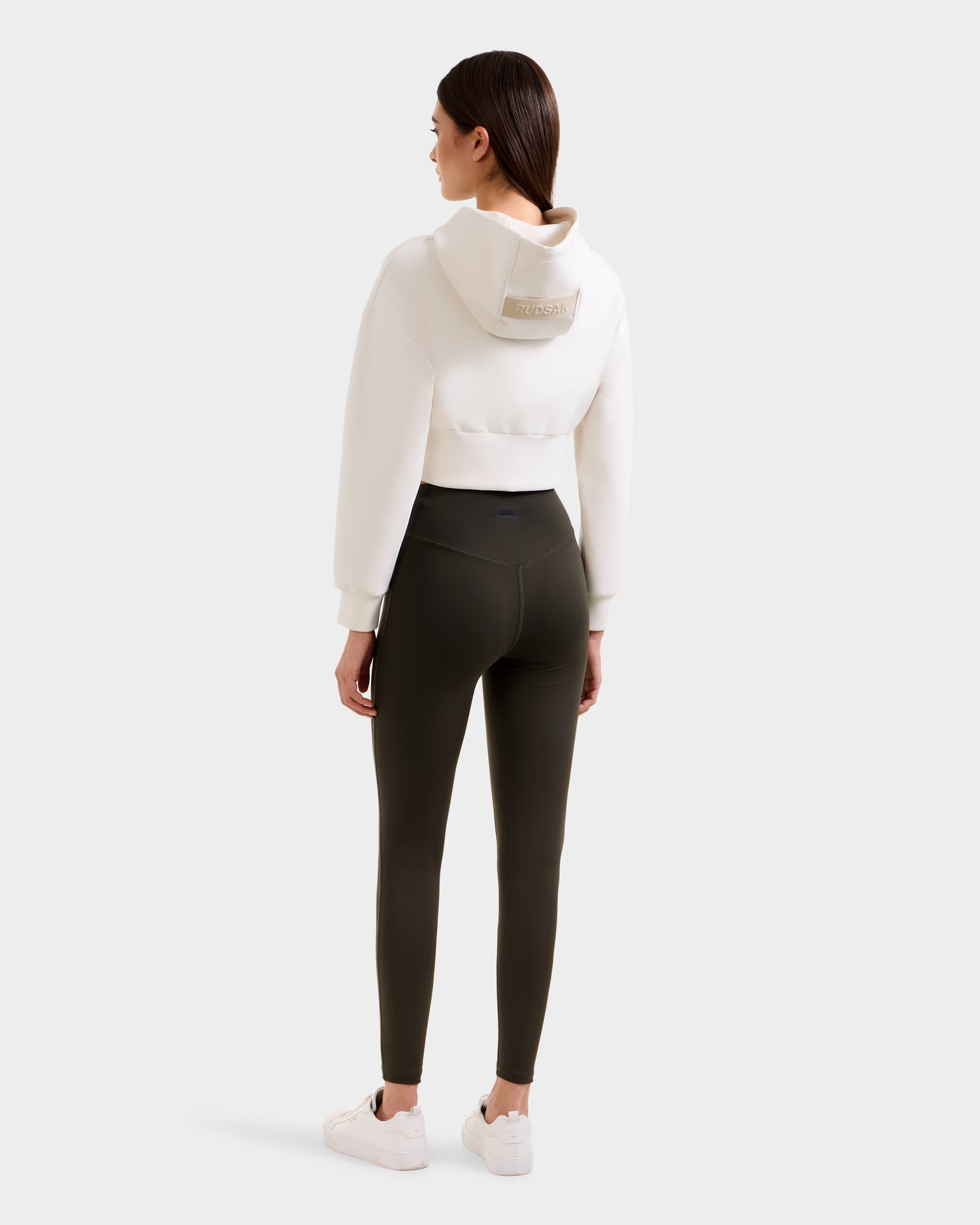 A woman faces away, wearing a cropped white hoodie with a hood and high-waisted Rudsak SONOMA - 8525071 Dark Olive leggings, paired with white sneakers, against a plain light background.