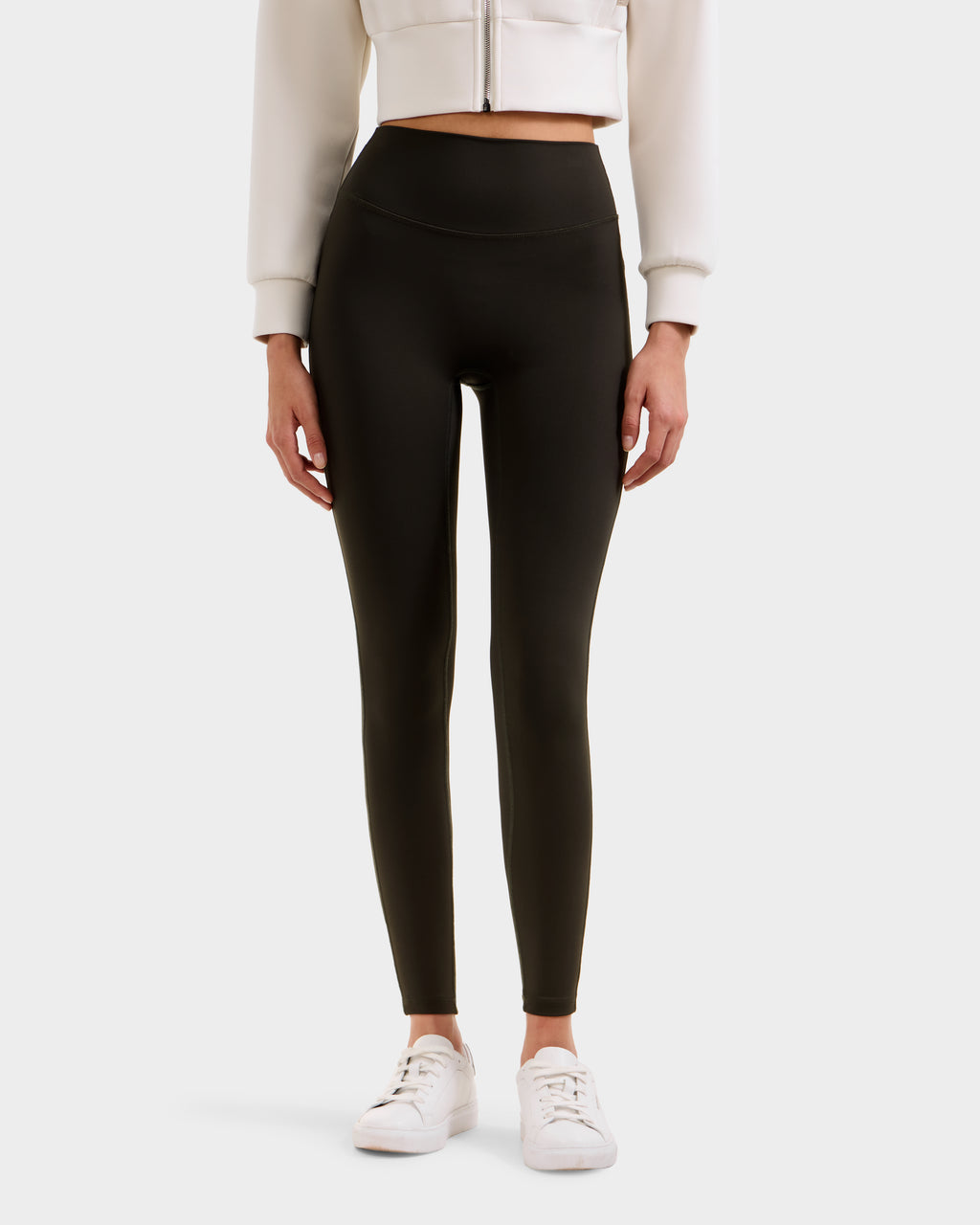 A person in high-waisted dark olive Rudsak SONOMA - 8525071 leggings, a cropped light jacket, and white sneakers stands against a plain background. The image is cropped from the shoulders down.