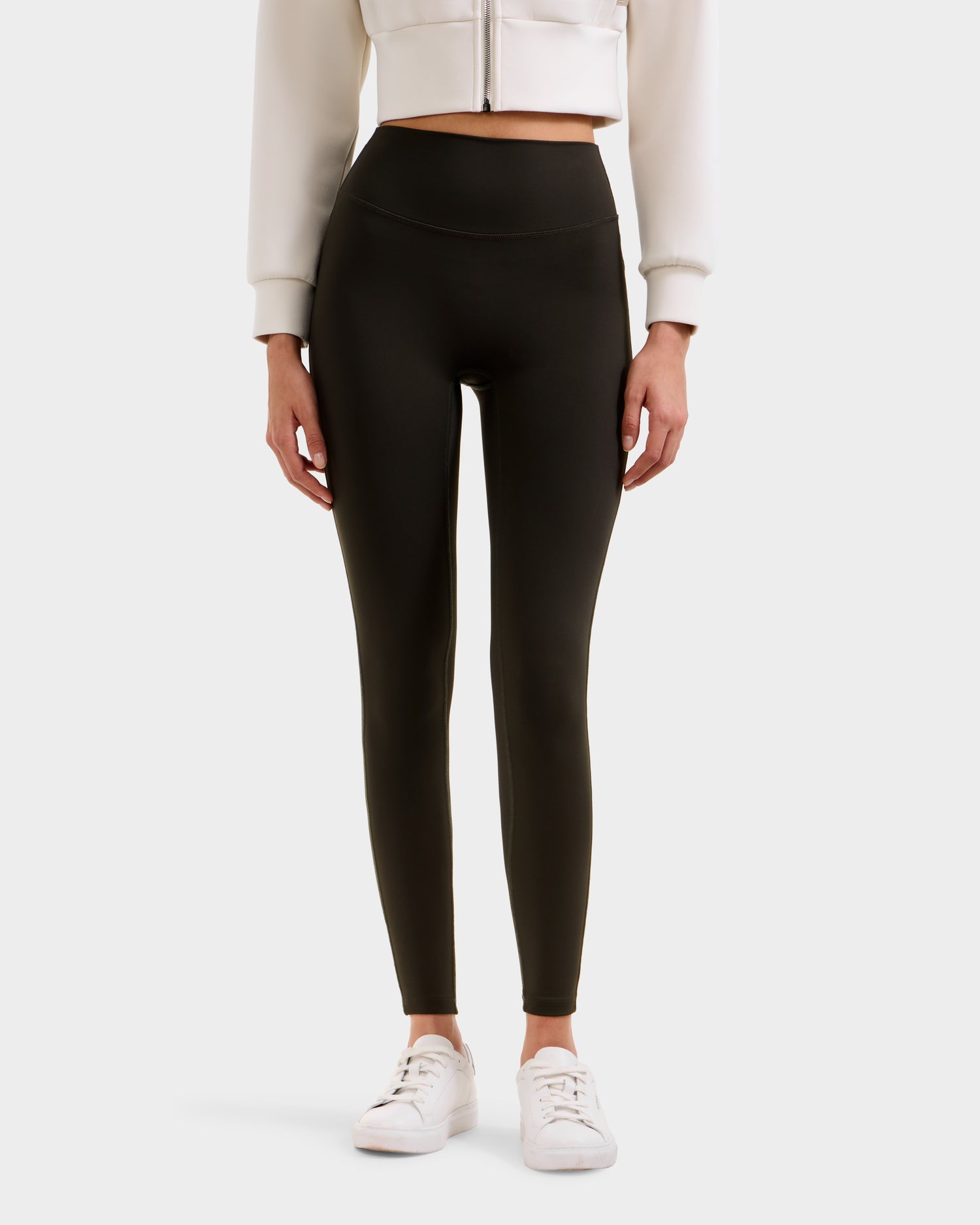 A person in high-waisted dark olive Rudsak SONOMA - 8525071 leggings, a cropped light jacket, and white sneakers stands against a plain background. The image is cropped from the shoulders down.