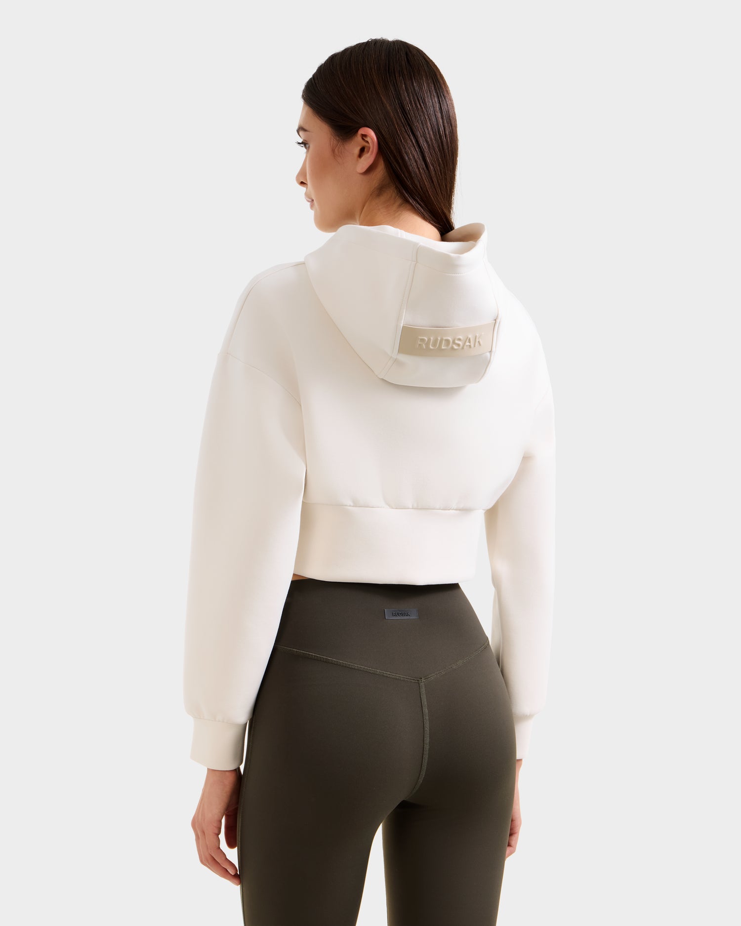 A woman with straight brown hair wears a cream cropped Rudsak hoodie with logo on the hood and high-waisted SONOMA - 8525071 Dark Olive leggings. She stands facing away, highlighting the back of her outfit.