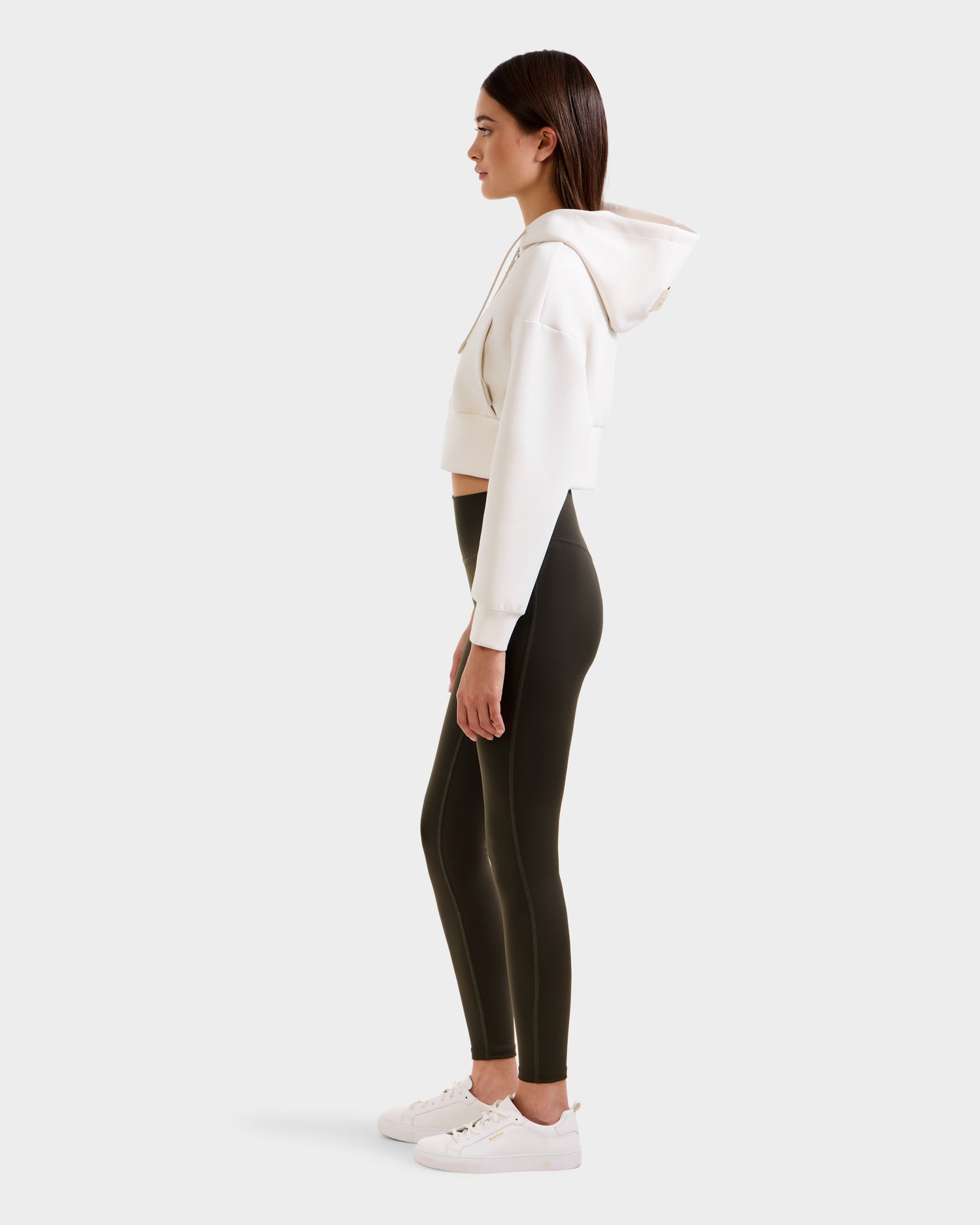 A woman stands in profile wearing a cropped white hoodie, Rudsak SONOMA - 8525071 Dark Olive versatile workout leggings, and white sneakers against a plain light background.