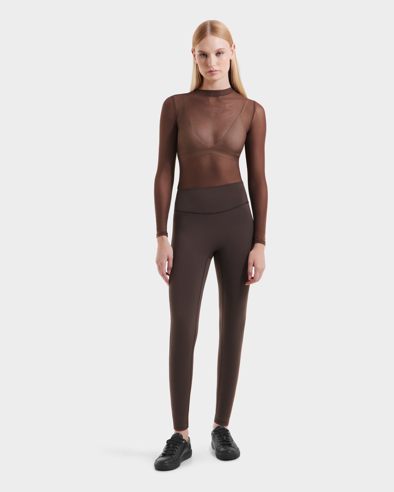 A woman with straight blonde hair wears a sheer brown long-sleeve top over a bralette, paired with Rudsak SONOMA - 8525071 Espresso high-performance leggings and black sneakers, standing against a plain white background.