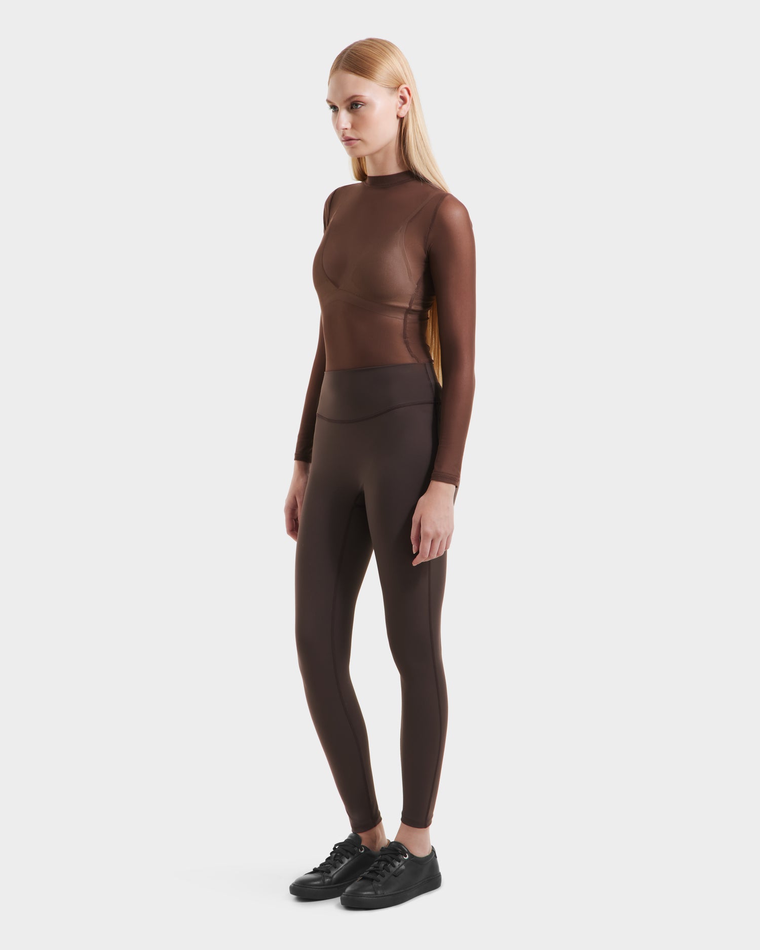 A woman with straight blonde hair stands against a white background, wearing a fitted brown long-sleeve top, Rudsak’s SONOMA - 8525071 Espresso high-performance leggings, and black sneakers. She is facing slightly to the side.