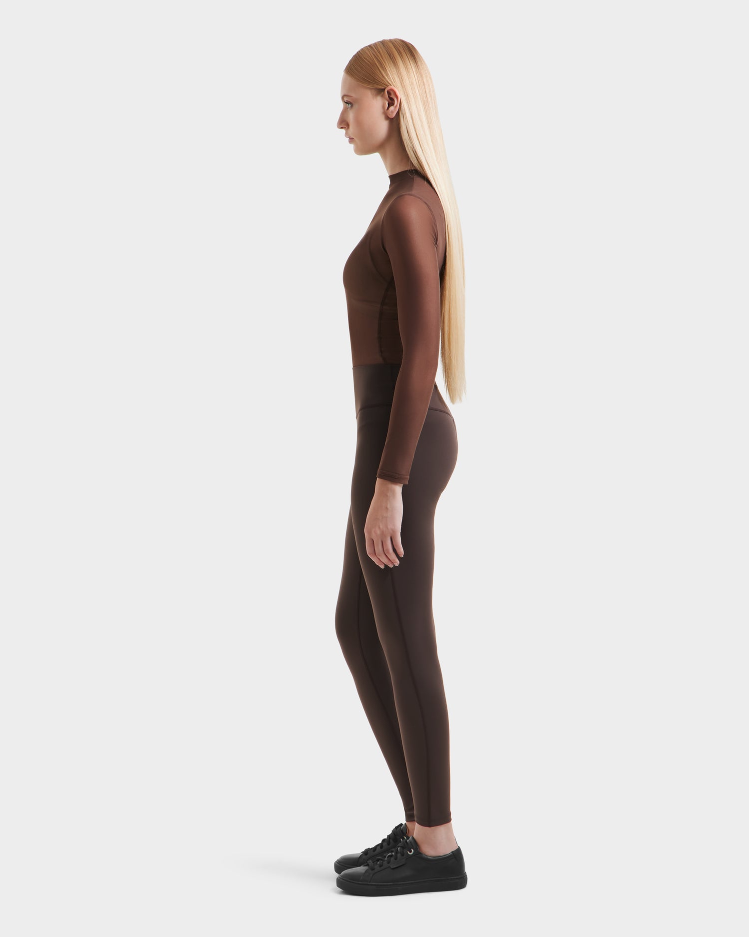 A woman with long blonde hair stands in profile, wearing a fitted brown long-sleeve top and Rudsak SONOMA - 8525071 Espresso leggings with black sneakers, set against a plain white background.