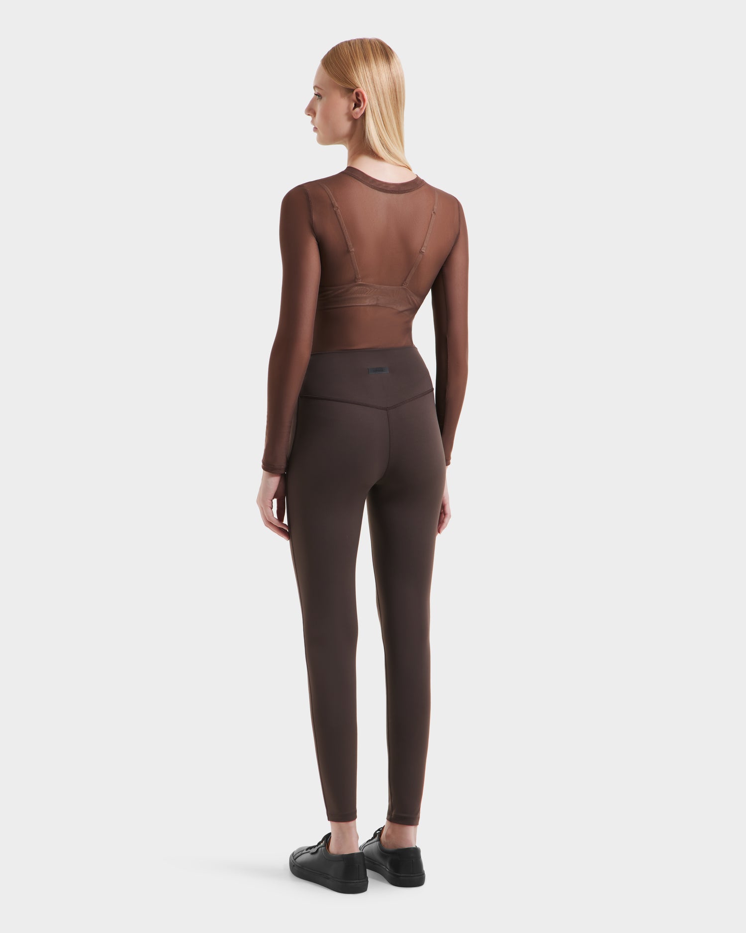 A woman with straight blonde hair faces away, wearing a sheer brown top, brown bralette, Rudsak’s SONOMA - 8525071 Espresso leggings, and black shoes against a plain white background.