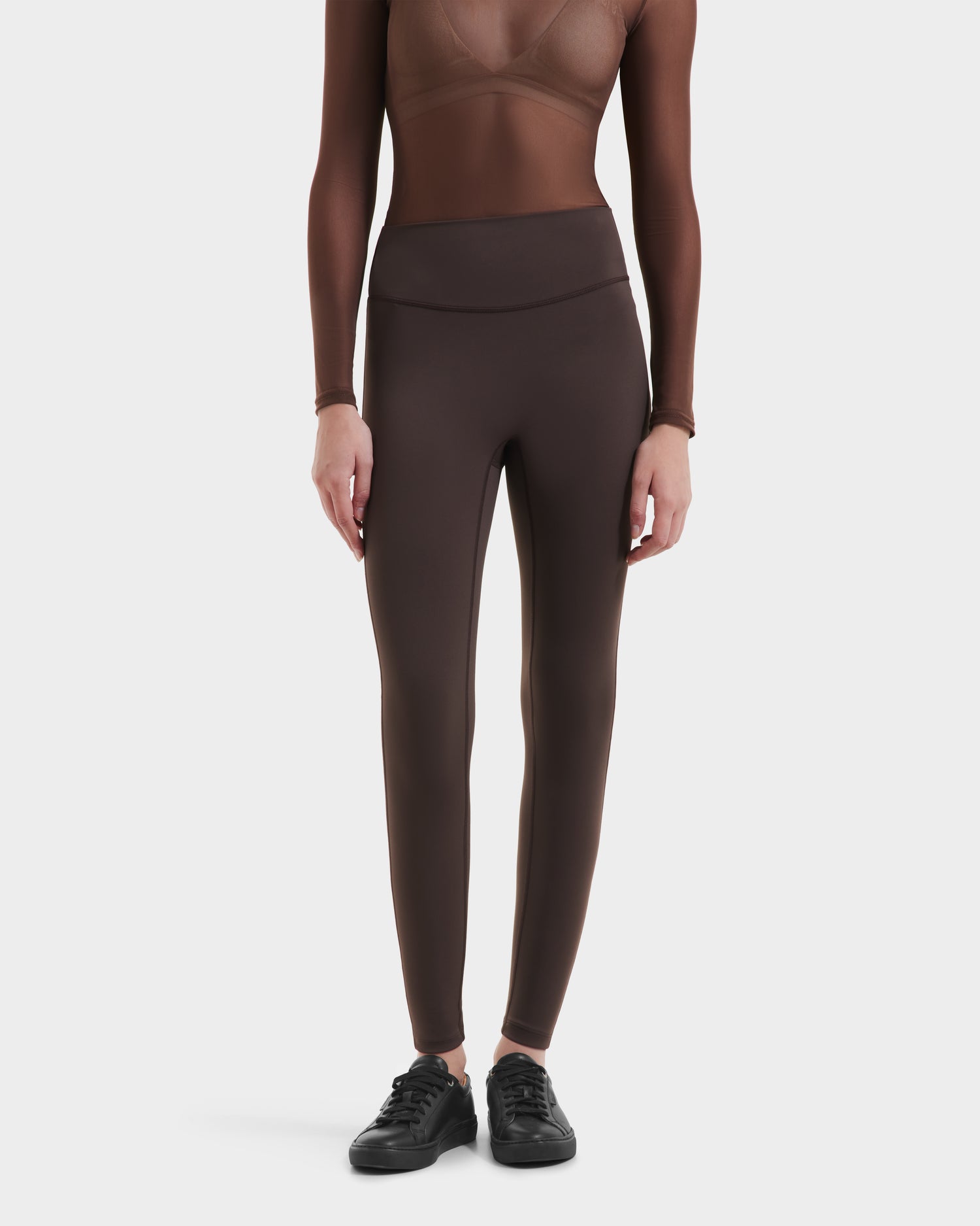A person wearing a sheer brown long-sleeve top and Rudsak’s SONOMA - 8525071 Espresso leggings with black sneakers stands against a plain white background, highlighting versatile activewear for any occasion.