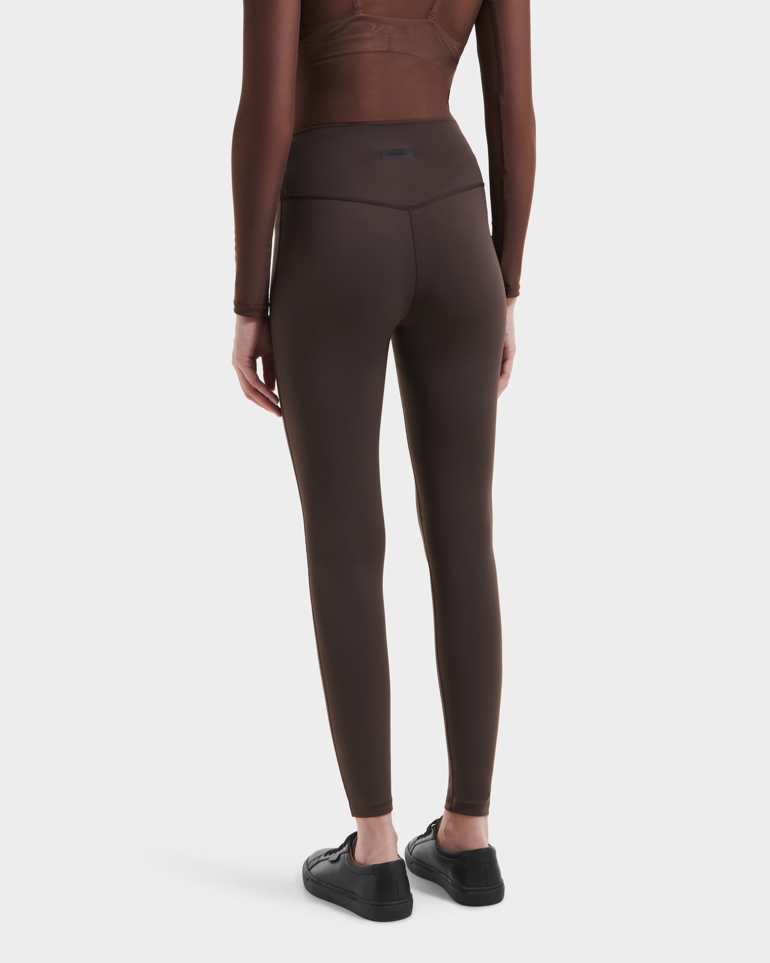 A person wearing a fitted brown long-sleeve top, Rudsak SONOMA - 8525071 Espresso high-performance leggings, and black sneakers stands with their back to the camera against a white background.