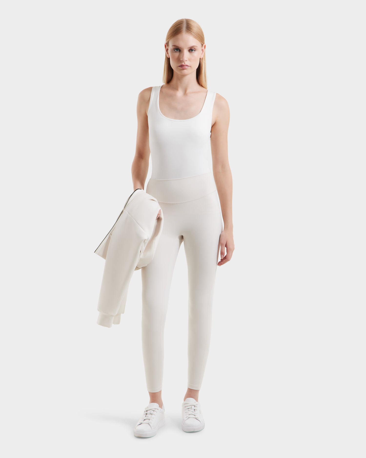 A woman with straight blonde hair wears a sleeveless white top, high-waisted Rudsak SONOMA - 8525071 Vanilla leggings, and white sneakers. Holding a light jacket in her left hand, she stands against a plain white background.
