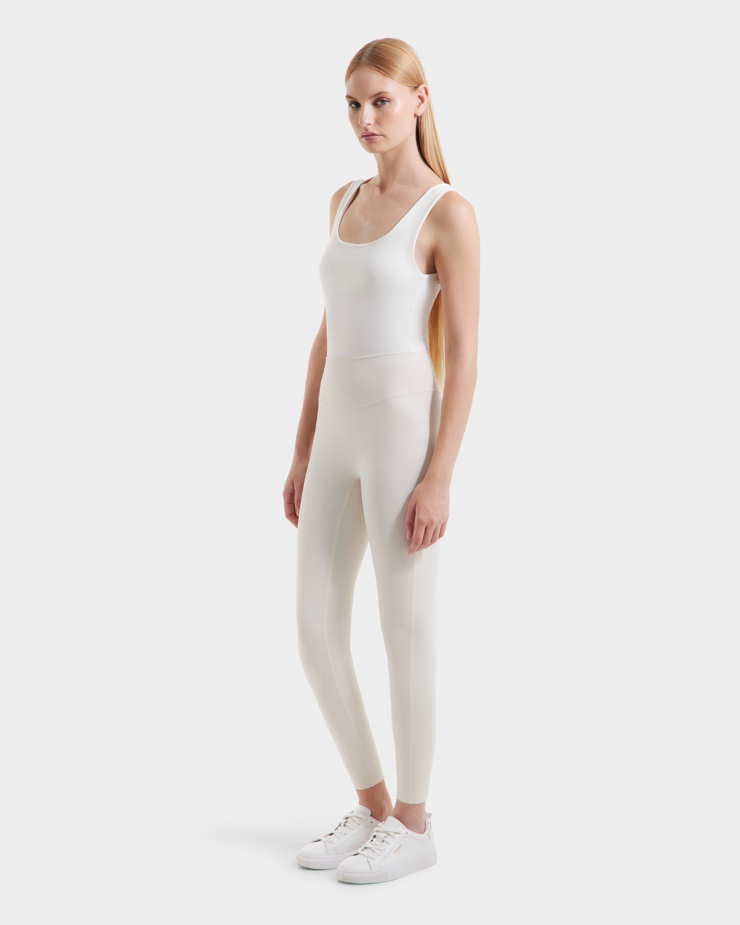 A woman with long blonde hair, facing slightly left, wears a fitted white tank top, Rudsak SONOMA - 8525071 Vanilla high-performance leggings, and white sneakers against a plain white background.