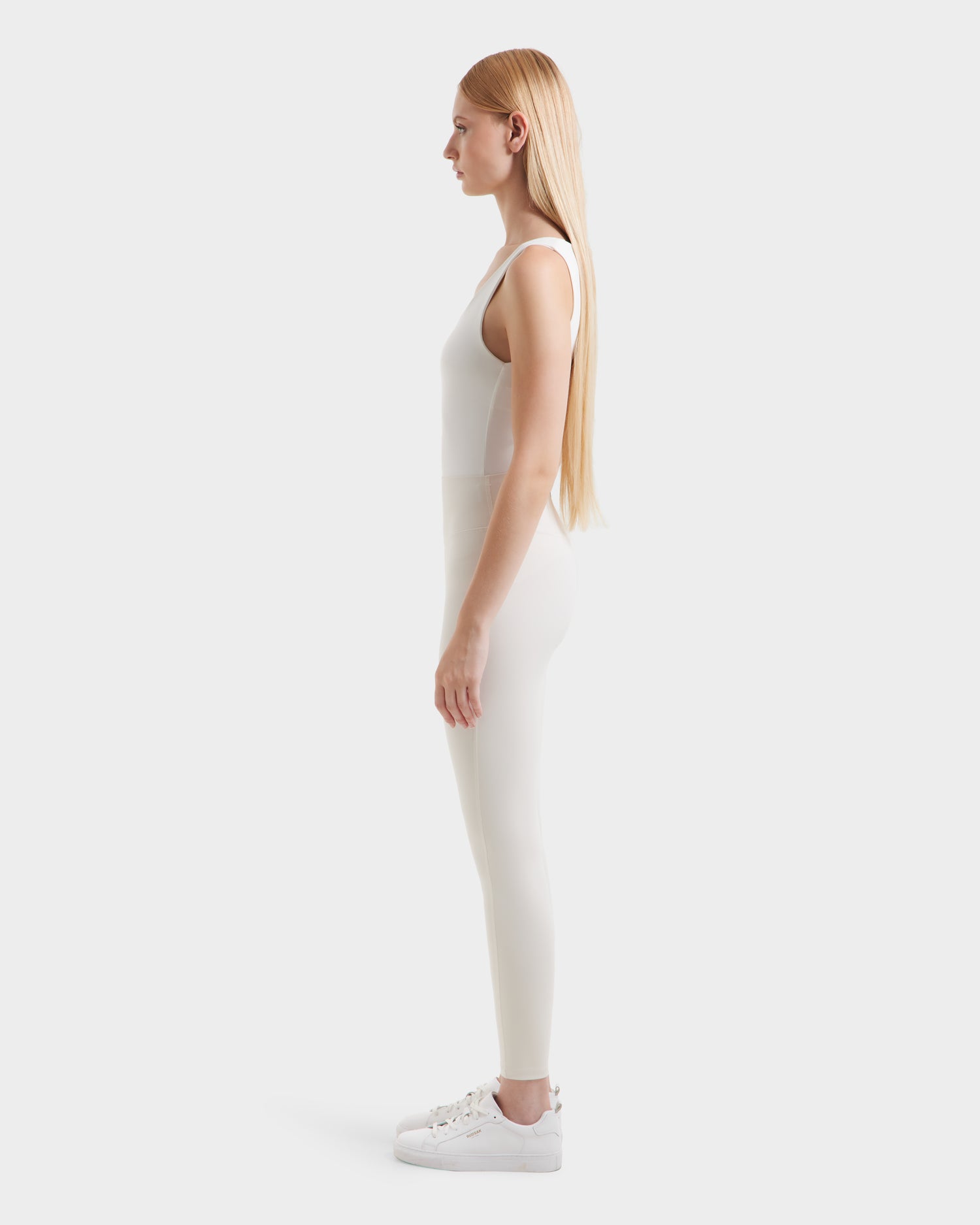 A woman with long straight blonde hair stands in profile, facing left. She wears a fitted white sleeveless top, Rudsak SONOMA - 8525071 Vanilla leggings, and white sneakers against a plain light gray background.