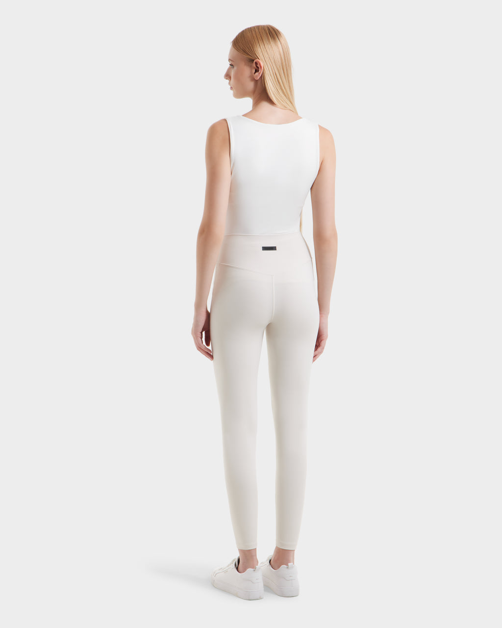 A woman with straight blonde hair, facing away, wears a sleeveless white top, high-waisted Rudsak SONOMA - 8525071 Vanilla leggings, and white sneakers against a plain light background.