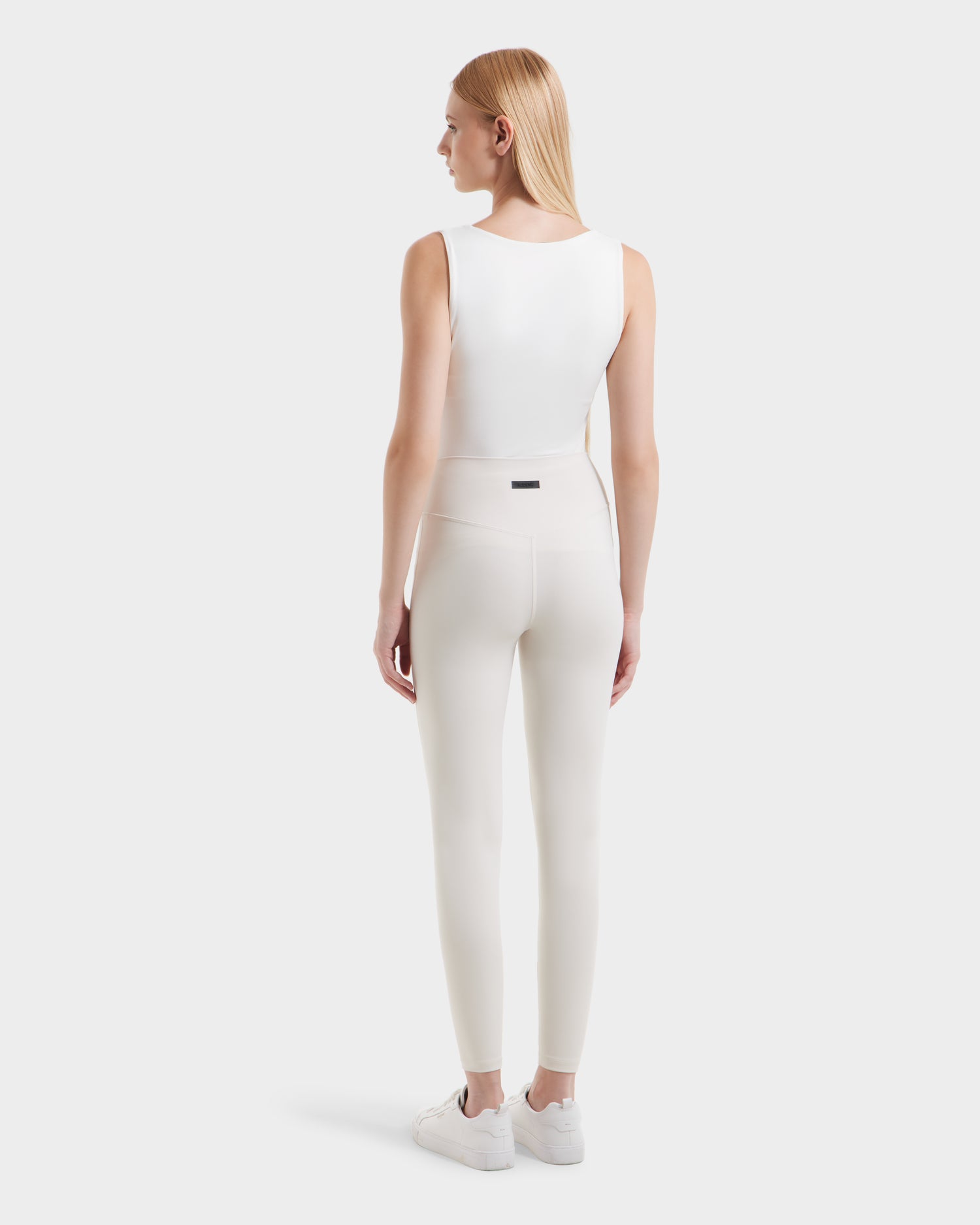 A woman with straight blonde hair, facing away, wears a sleeveless white top, high-waisted Rudsak SONOMA - 8525071 Vanilla leggings, and white sneakers against a plain light background.