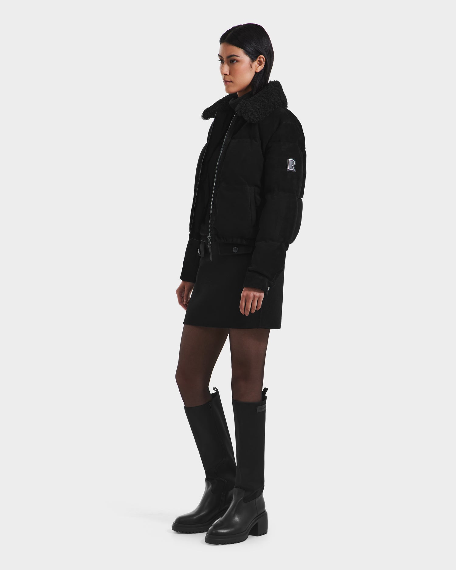 A person with short dark hair wears the Rudsak STACEY - 8125049 Black women’s aviator jacket with a removable shearling collar, paired with a black skirt, sheer black tights, and tall black boots against a plain light background.