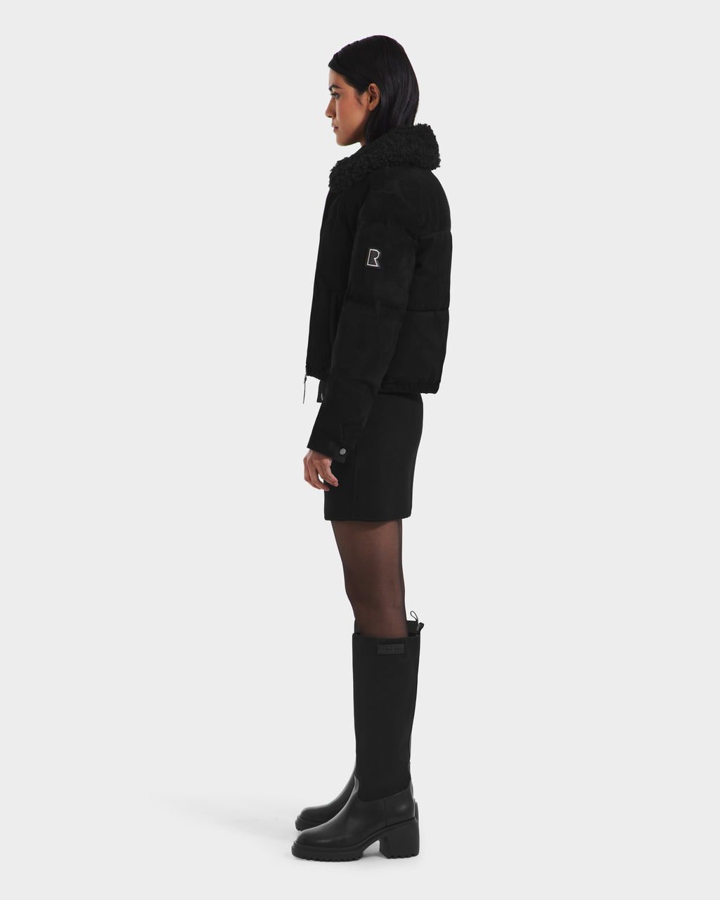 A woman stands in profile against a white background, wearing the Rudsak STACEY - 8125049 Black aviator jacket with removable shearling collar, paired with a black mini skirt and knee-high boots. Her hair is down and hands at her sides.
