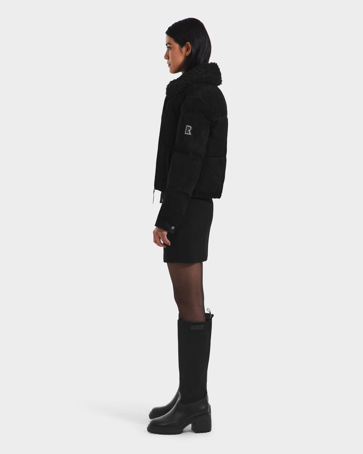 A woman stands in profile against a white background, wearing the Rudsak STACEY - 8125049 Black aviator jacket with removable shearling collar, paired with a black mini skirt and knee-high boots. Her hair is down and hands at her sides.