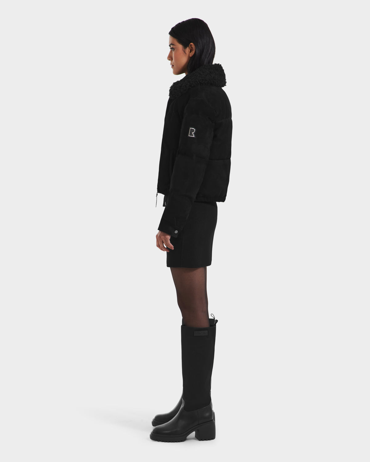 A woman stands in profile against a white background, wearing the Rudsak STACEY - 8125049 Black aviator jacket with removable shearling collar, paired with a black mini skirt and knee-high boots. Her hair is down and hands at her sides.