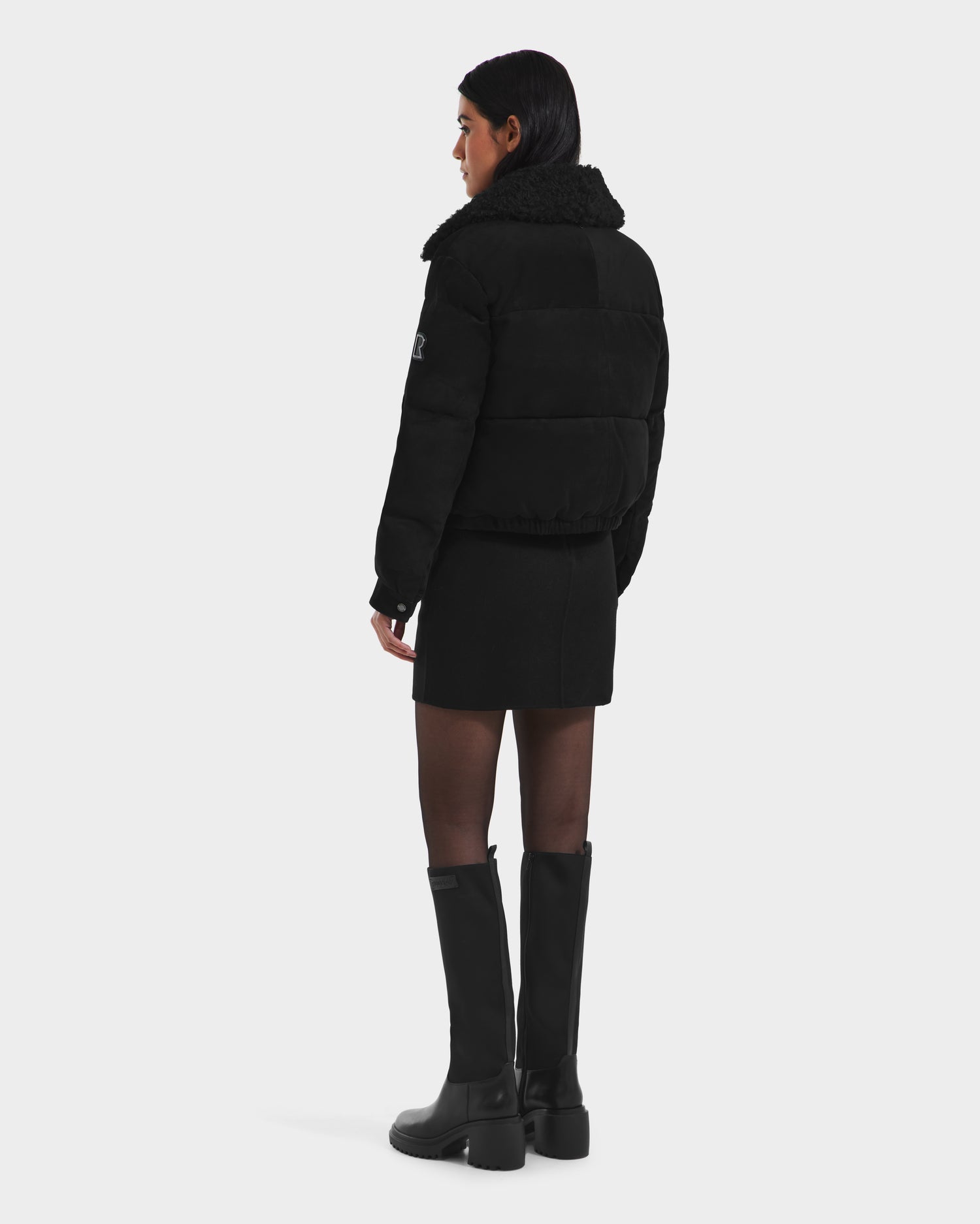A woman faces away, wearing the Rudsak STACEY - 8125049 Black women’s aviator jacket with a removable shearling collar, paired with a black mini skirt, knee-high boots, and sheer tights against a plain white background.
