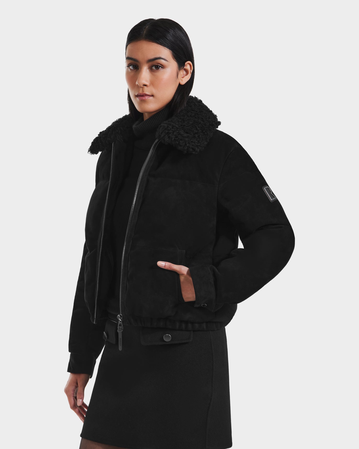 A woman wears the Rudsak STACEY - 8125049 Black women’s aviator jacket with a shearling collar over a black turtleneck and skirt, standing against a plain white background.